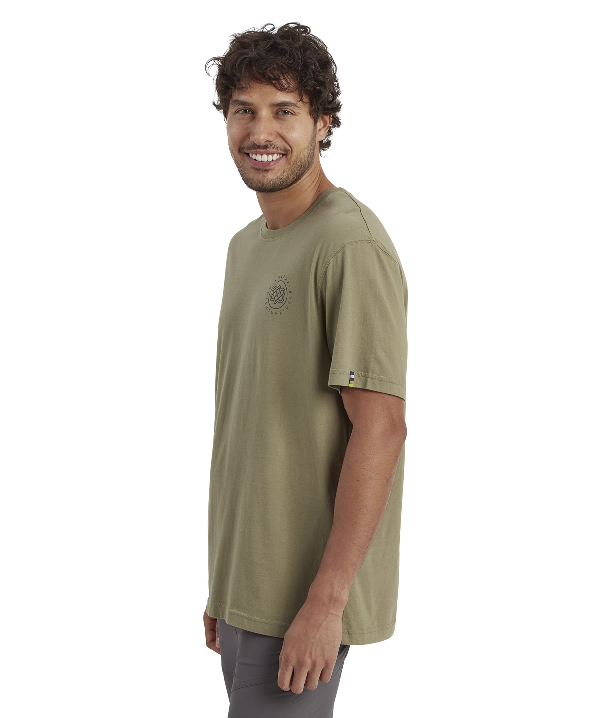 The full-length view of the same man wearing the Sherpa Adventure Gear Summit T-Shirt in Green, paired with grey hiking pants and mustard-yellow hiking shoes. He stands in a relaxed pose, showcasing the casual outdoor style of the outfit.