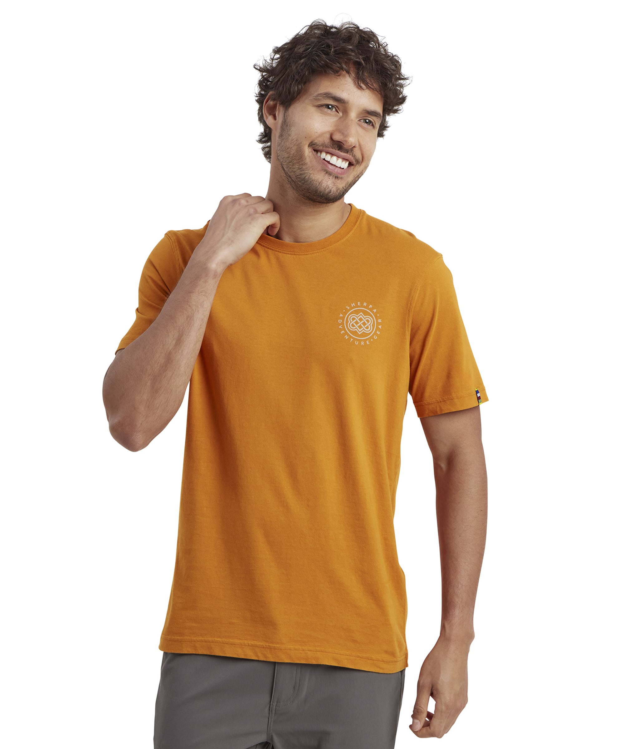 A man with curly brown hair and light stubble smiles while wearing an Sherpa Adventure Gear Summit T-Shirt in Orange with a crew neck. The front features a small circular Sherpa Adventure Gear logo on the left chest area. He stands with his left hand in his pocket and his right hand resting on his neck, wearing dark grey trousers. The fabric looks soft and lightweight.