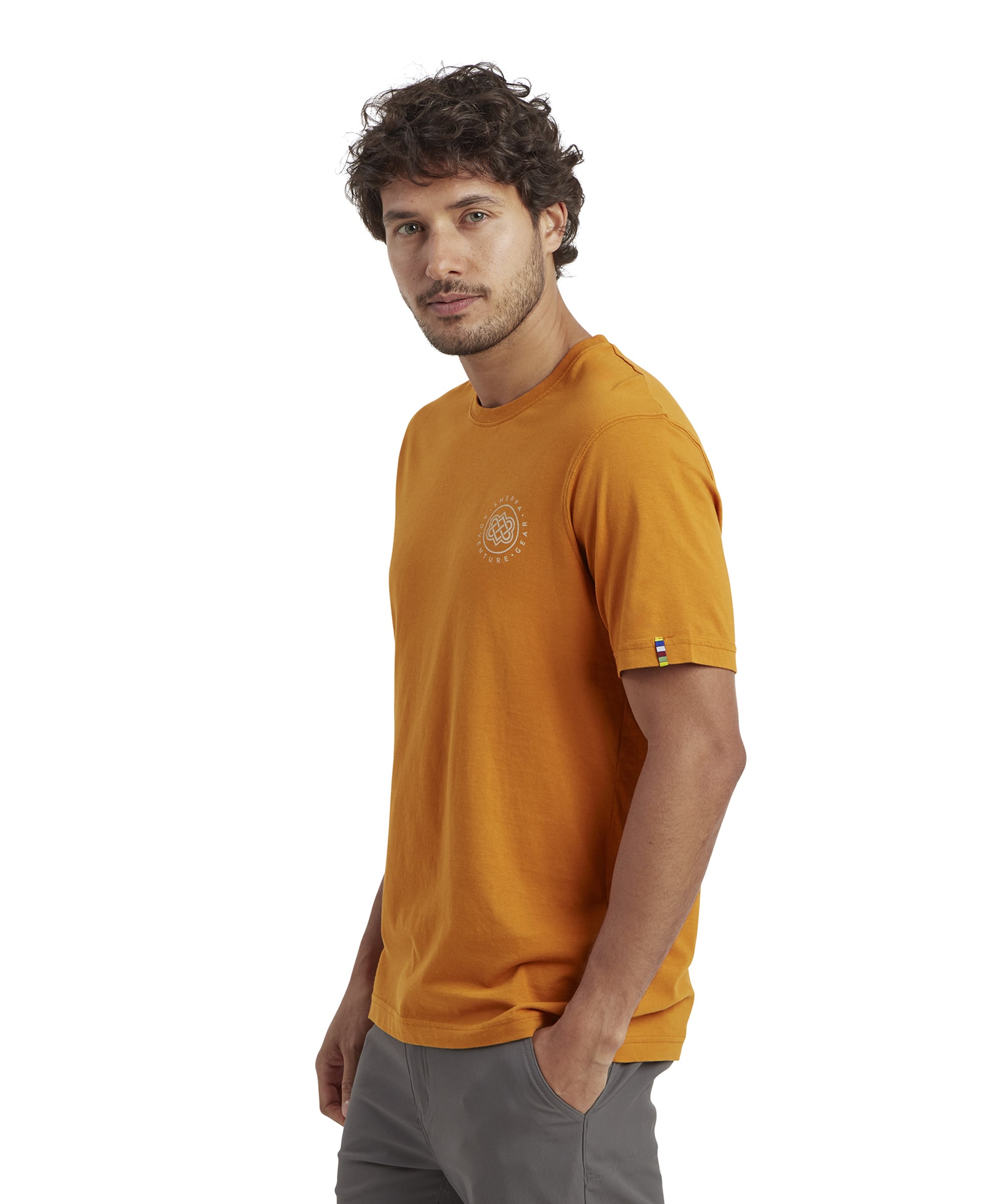 A profile view of the man wearing the Sherpa Adventure Gear Summit T-Shirt in Orange. The left sleeve has a small woven label with a multicolored stripe, adding a subtle detail. The shirt fits snugly on the shoulders and arms, and the hem sits naturally at the hips.