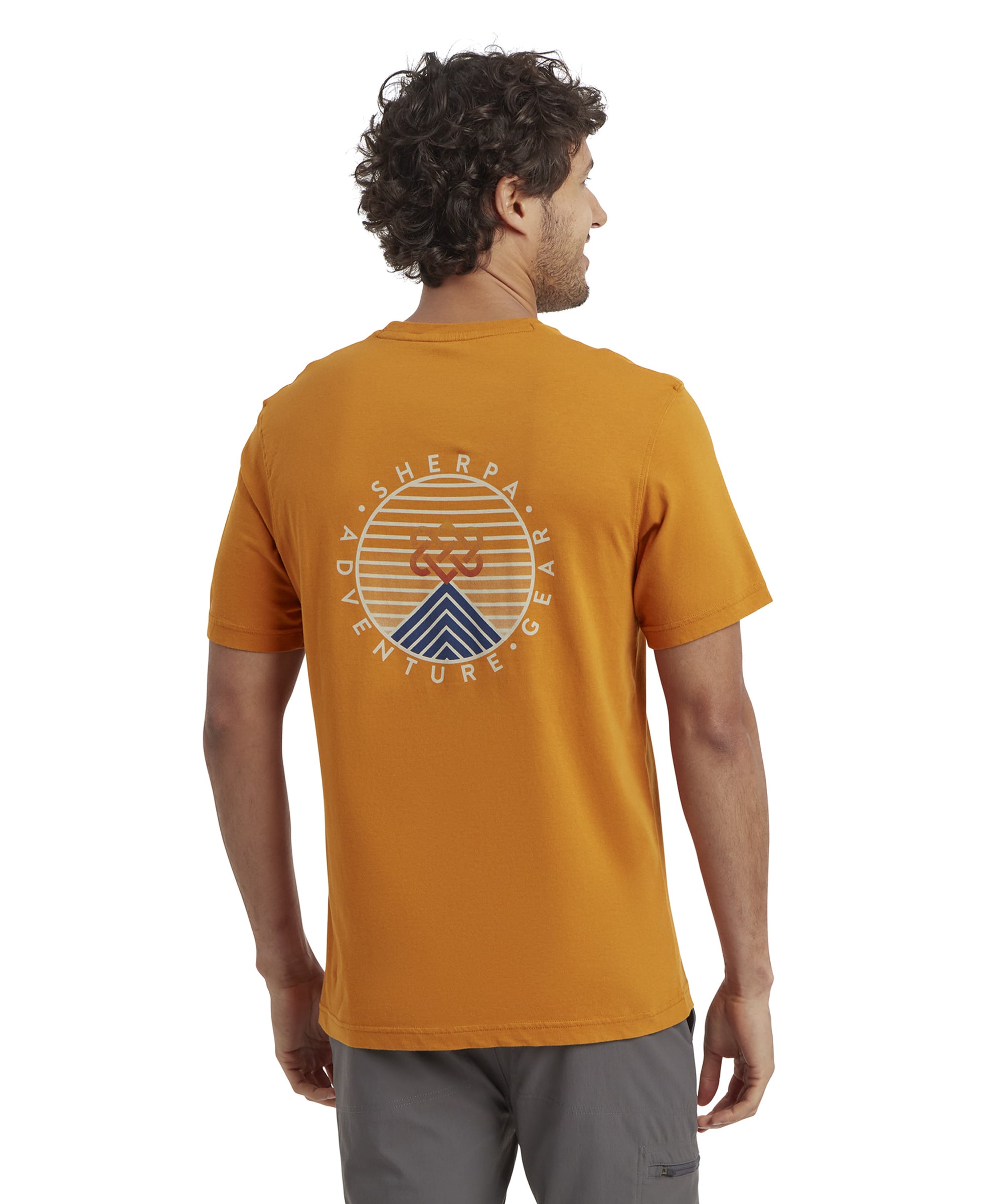 The back of the Sherpa Adventure Gear Summit T-Shirt in Orange features a large Sherpa Adventure Gear graphic in the center. The design includes a circular shape with horizontal orange stripes, a blue geometric mountain peak at the bottom, and the Sherpa Adventure Gear logo in the middle. The model stands with his back to the camera, slightly turned to show the full design.