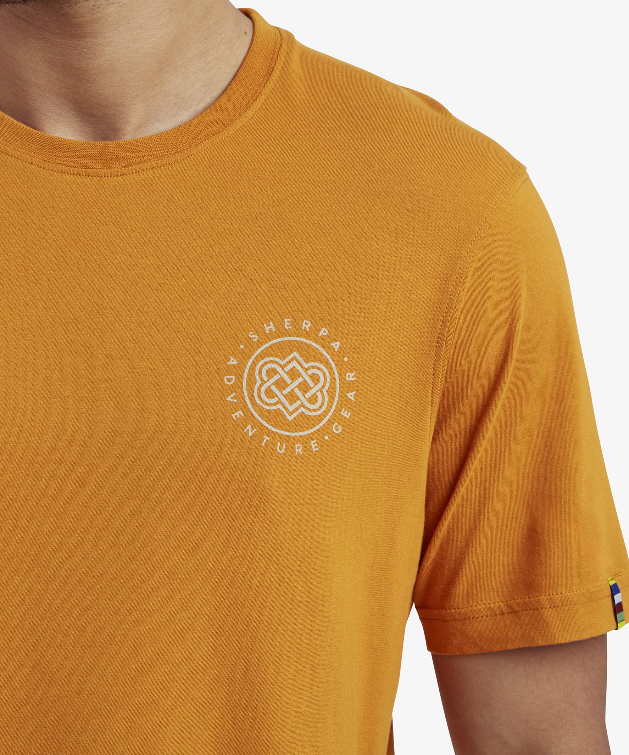 A detailed shot of the small white circular Sherpa Adventure Gear logo on the left chest area on the Sherpa Adventure Gear Summit T-Shirt in Orange. The logo includes a stylized knot design surrounded by the words "Sherpa Adventure Gear."