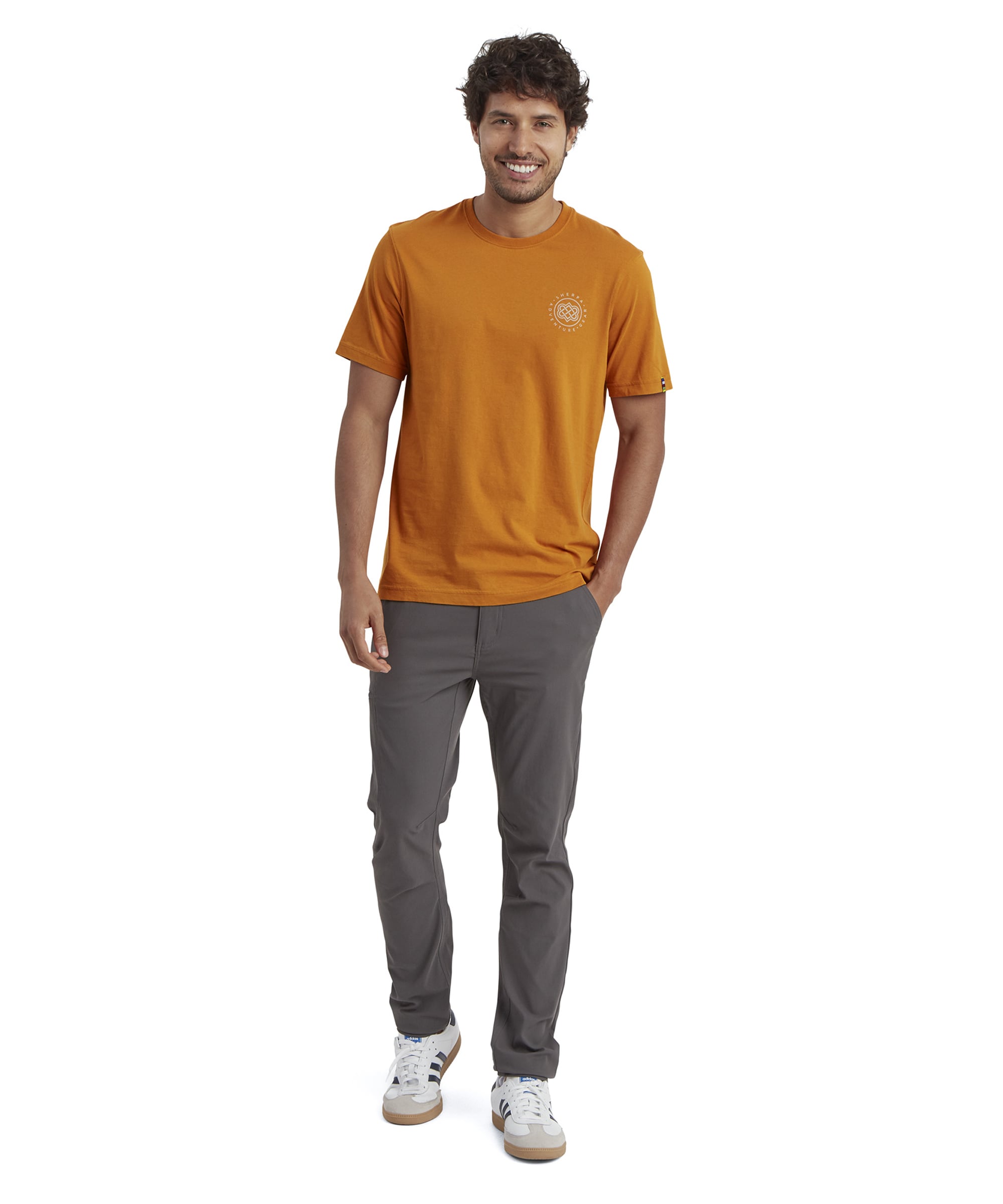 The same model stands casually in the Sherpa Adventure Gear Summit T-Shirt in Orange and dark grey trousers, wearing white trainers. His hands are in his pockets, and he looks straight at the camera with a relaxed expression. The t-shirt falls to the hips, providing a comfortable fit.