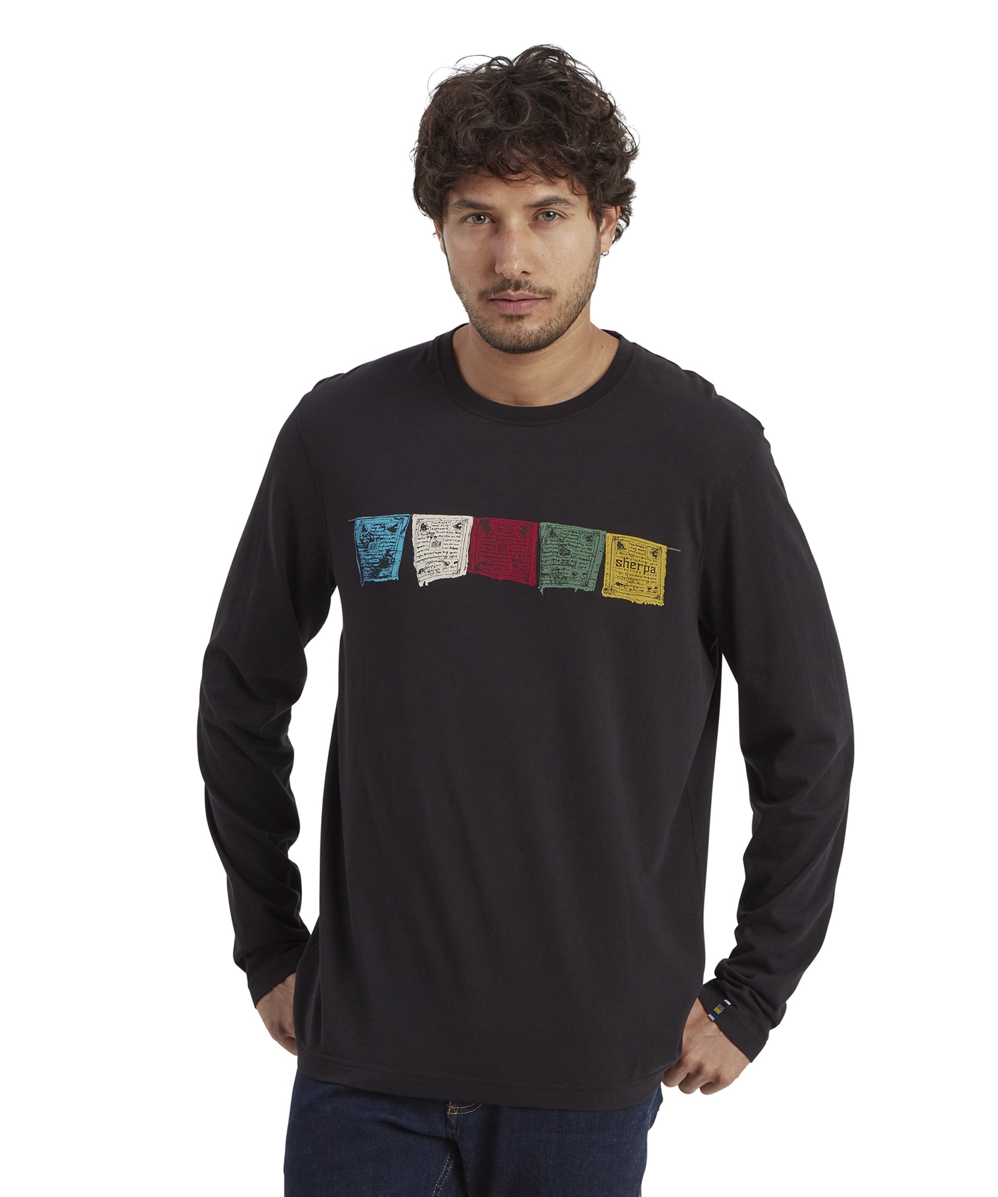 A male model wearing a Sherpa Adventure Gear Tarcho Long Sleeve T-Shirt in Black featuring a row of colorful Tibetan prayer flags printed across the chest in blue, white, red, green, and yellow. The relaxed fit of the shirt is paired with dark jeans, creating a casual look.