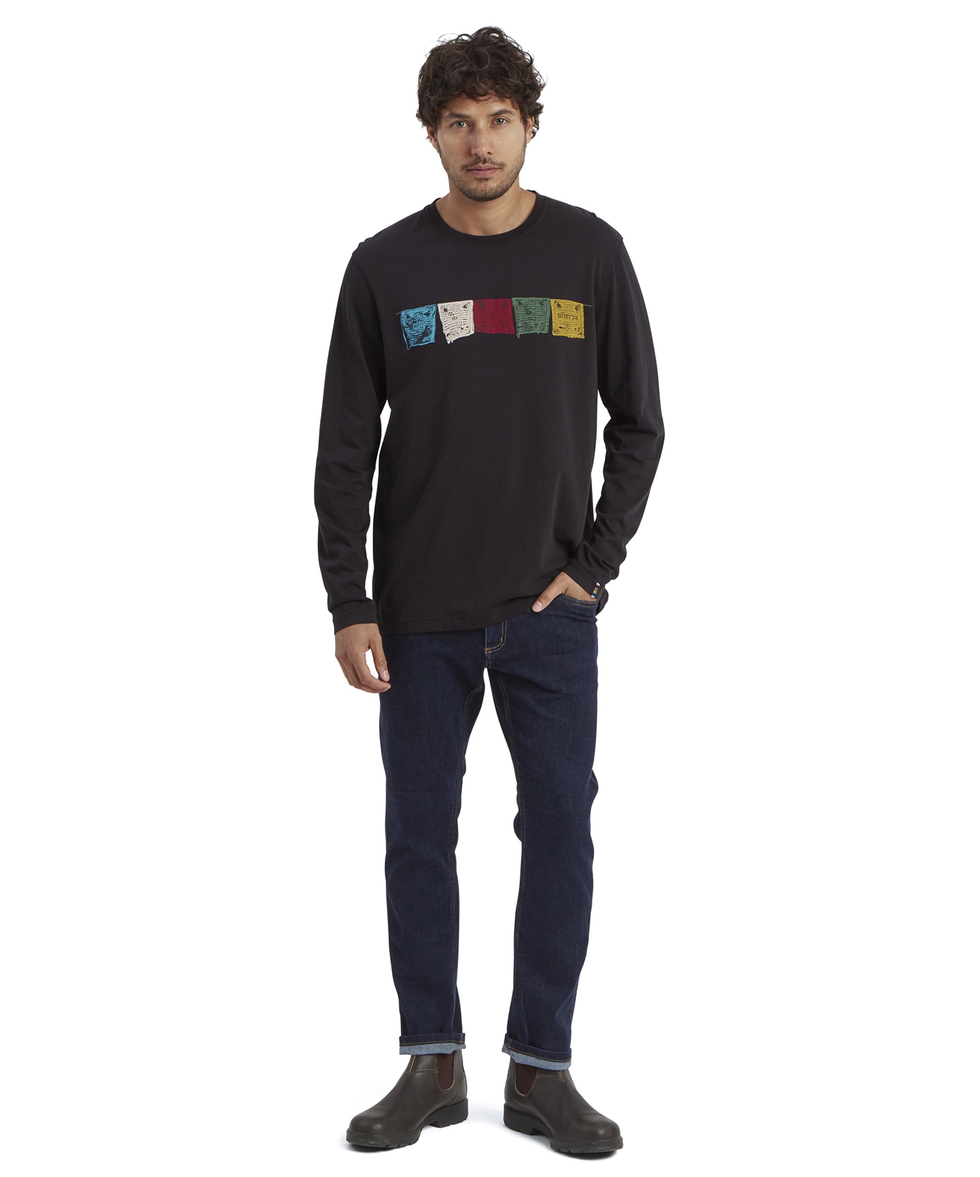 Full-body view of the model in the Sherpa Adventure Gear Tarcho Long Sleeve T-Shirt in Black. The outfit is styled with dark jeans and brown leather boots. The colorful prayer flags on the t-shirt contrast against the black fabric, making the design stand out.