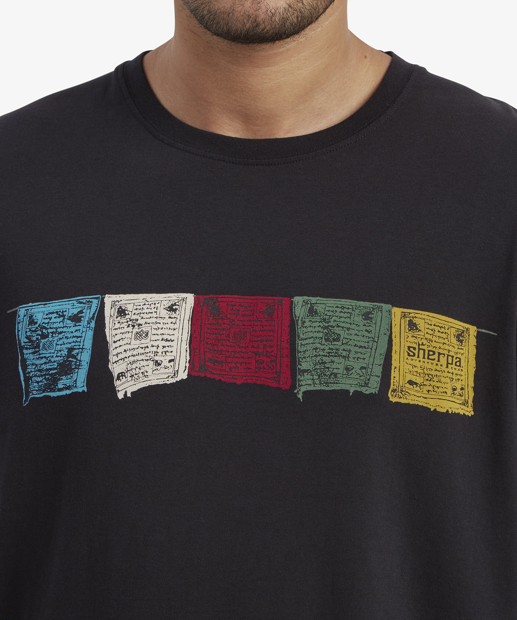 Close-up of the chest area, focusing on the vibrant prayer flag design. Each flag features intricate details of traditional Tibetan script and symbols, adding cultural significance and visual appeal to the Sherpa Adventure Gear Tarcho Long Sleeve T-Shirt in Black. The fabric appears soft and breathable.