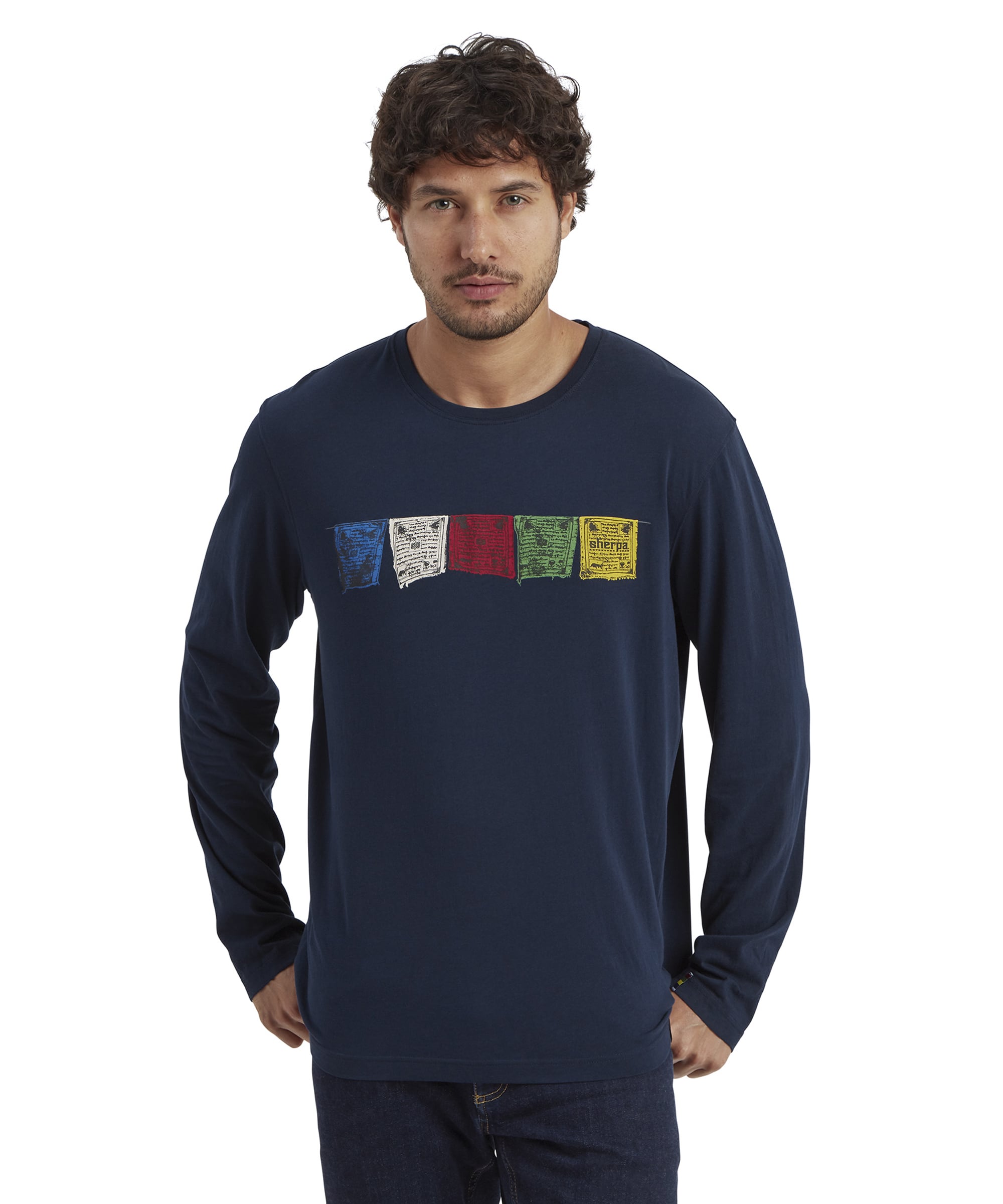 A man is wearing a Sherpa Adventure Gear Tarcho Long Sleeve T-Shirt in Blue with colorful printed prayer flags across the chest. The shirt has a relaxed fit and is paired with dark blue jeans. The model has a neutral expression and is standing against a white background.