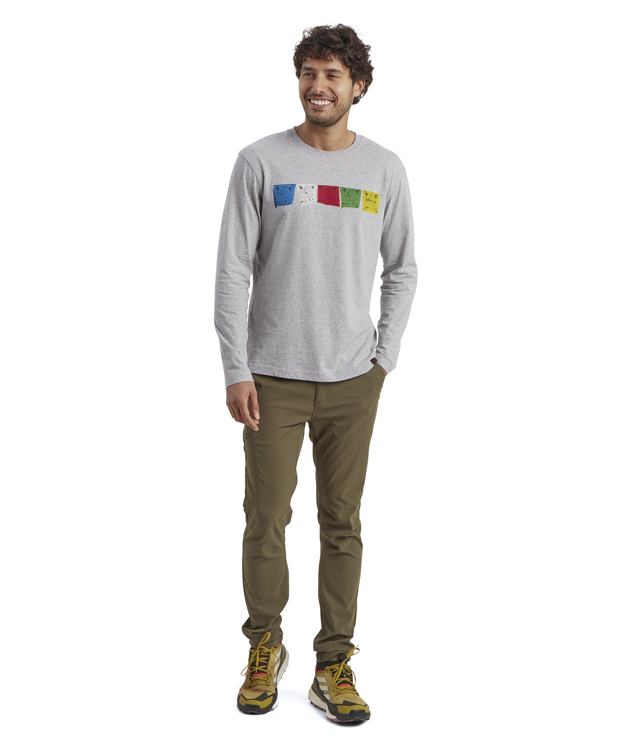 The model wears the Sherpa Adventure Gear Tarcho Long Sleeve T-Shirt in Grey paired with olive green trousers and yellow hiking trainers. The bright colors of the prayer flag graphic contrast against the muted grey shirt, while the model smiles, standing casually against a white background.