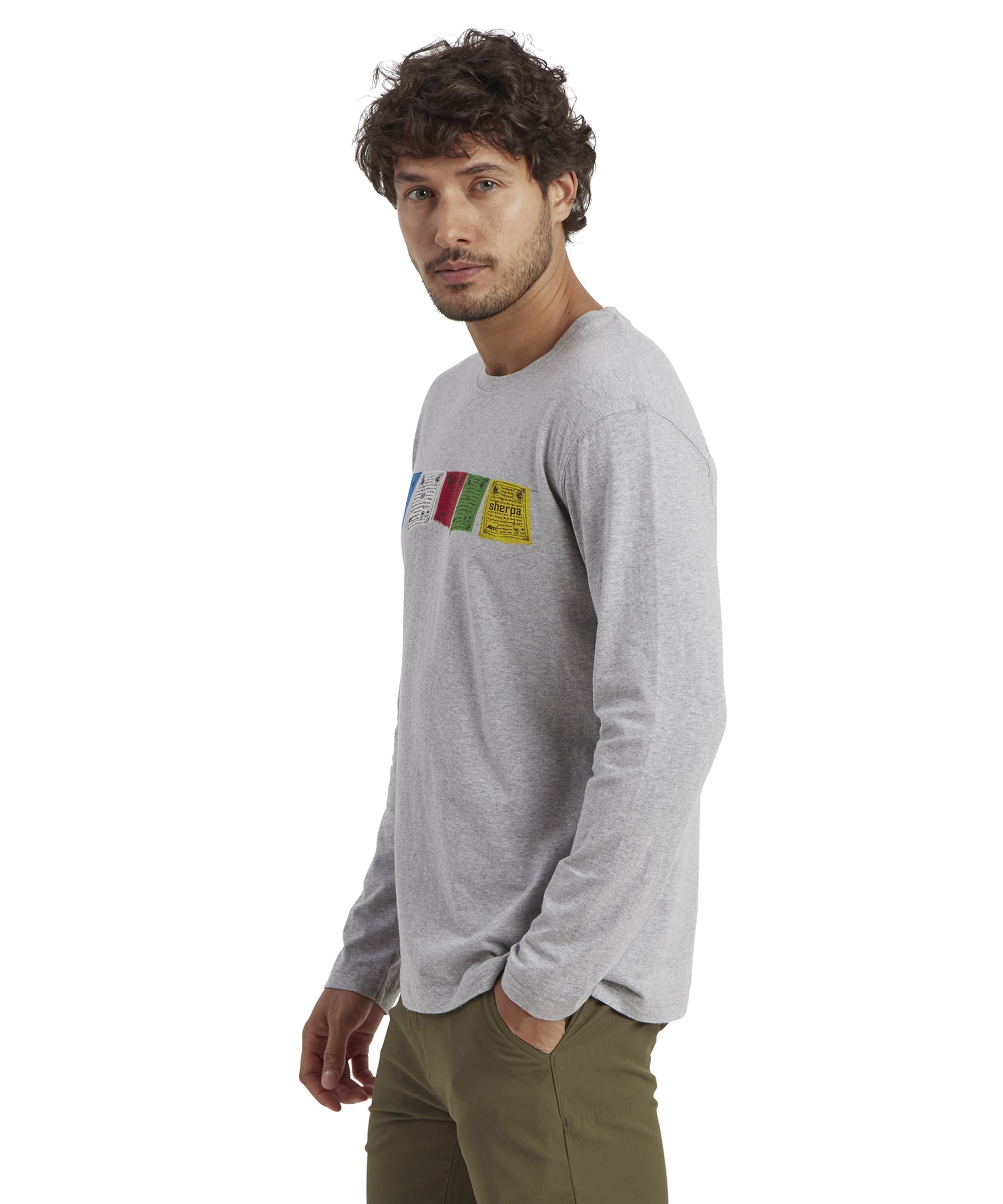 A side view showcases the fit and length of the Sherpa Adventure Gear Tarcho Long Sleeve T-Shirt in Grey, with the vibrant prayer flag graphics prominently displayed on the chest. The lightweight and breathable fabric complements the model's relaxed posture.