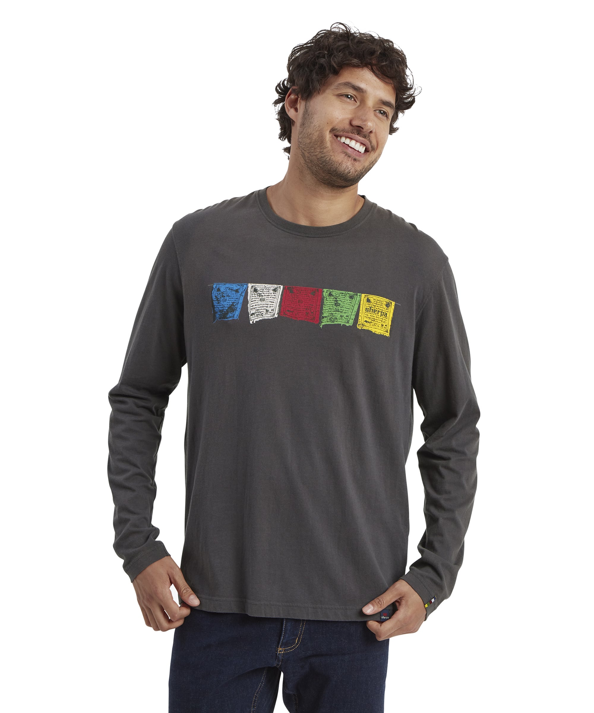 A male model with curly brown hair and a light beard is wearing a Sherpa Adventure Gear Tarcho Long Sleeve T-Shirt in Grey. The T-shirt features a horizontal row of colorful Tibetan prayer flag graphics in blue, white, red, green, and yellow across the chest. He is smiling, standing casually with his hands slightly in his front pockets, and looking to the side. He is also wearing dark jeans. The background is solid white.