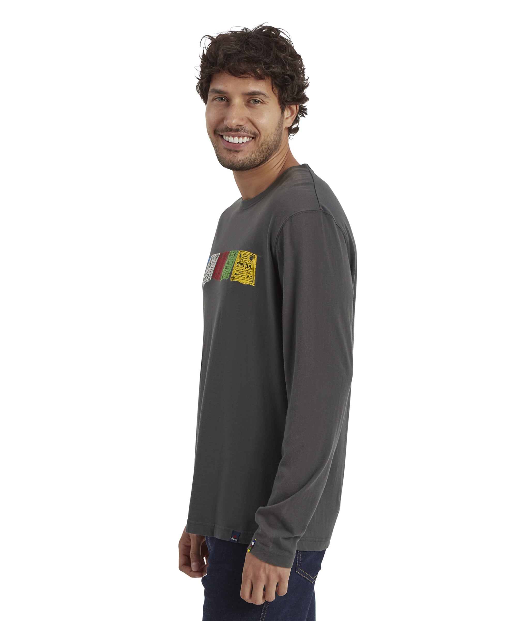 A profile shot of the male model highlights the side view of the Sherpa Adventure Gear Tarcho Long Sleeve T-Shirt in Grey. The colorful Tibetan prayer flag graphics are visible on the chest. He is smiling while standing casually, hands at his sides. The T-shirt fits comfortably, draping naturally over his torso. The plain white background contrasts with the T-shirt color.