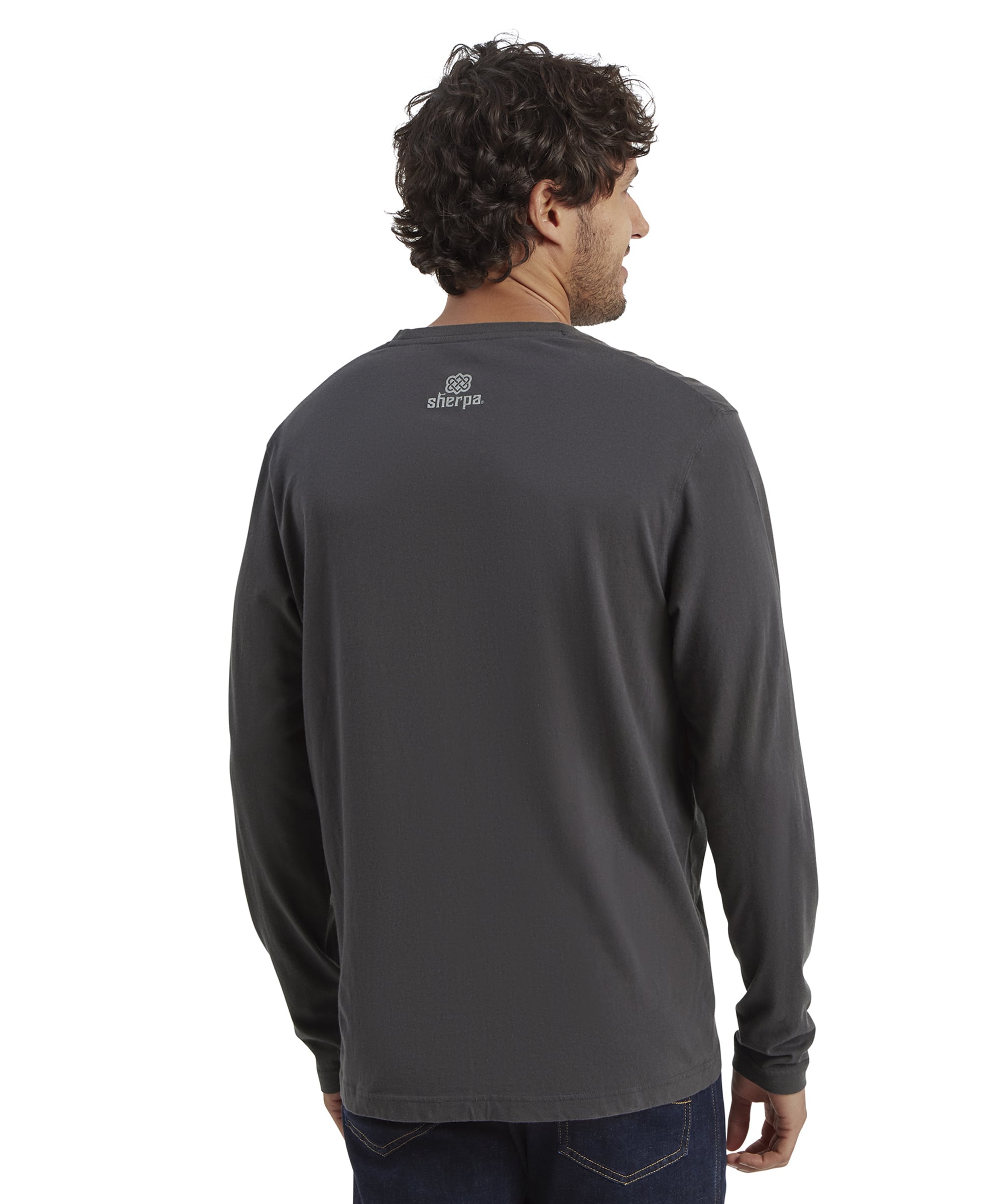 A back view of the male model shows the Sherpa Adventure Gear Tarcho Long Sleeve T-Shirt in Grey. The T-shirt features a small white Sherpa logo printed near the neckline on the back. The model stands straight with his head turned slightly to the side, revealing his profile. The T-shirt extends past his waist, covering the upper part of his jeans. The solid white background provides contrast.