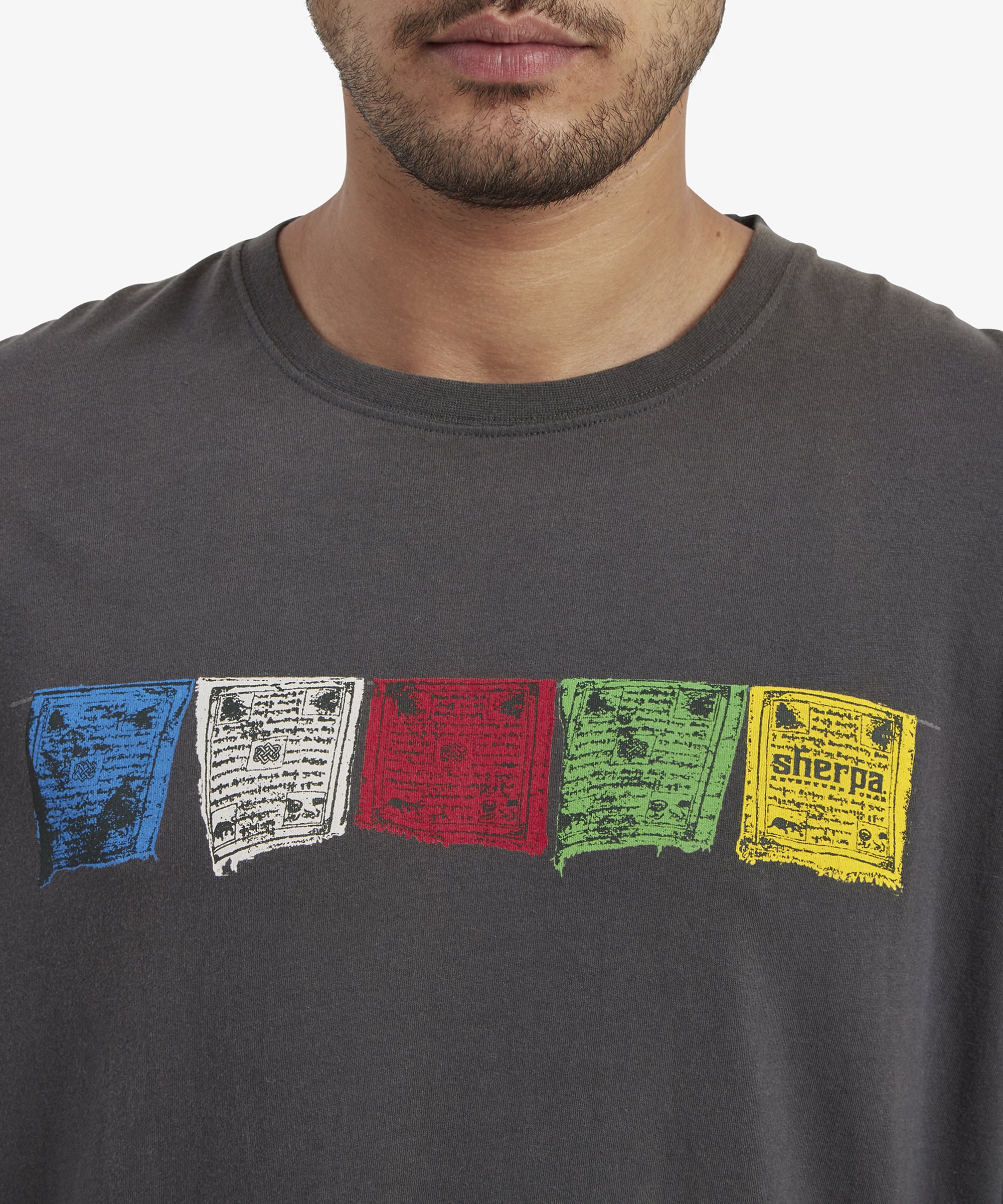 A close-up of the chest area of the Sherpa Adventure Gear Tarcho Long Sleeve T-Shirt in Grey focuses on the vibrant Tibetan prayer flag graphics. The design consists of five rectangular flags in blue, white, red, green, and yellow, each featuring intricate printed text and designs. The yellow flag prominently features the "Sherpa" logo. The close-up captures the fine details of the fabric texture and graphics.