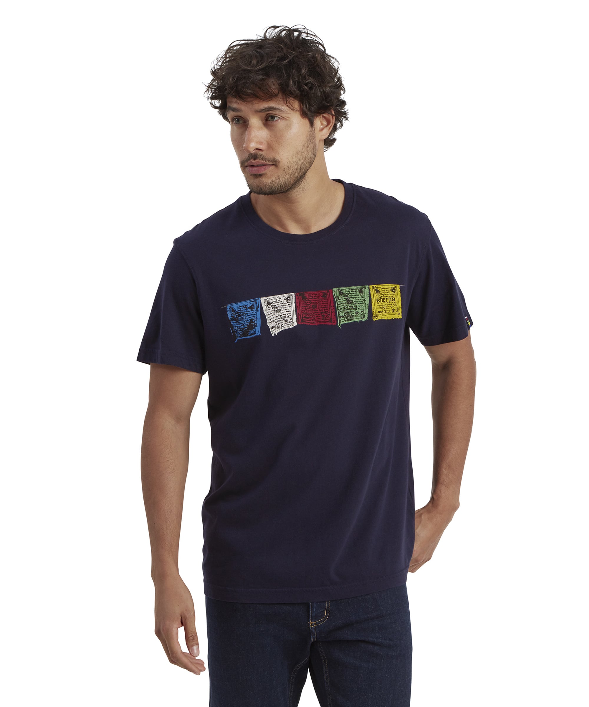 A Sherpa Adventure Gear Tarcho T-Shirt in Blue featuring colorful rectangular Tibetan prayer flag designs across the chest in blue, white, red, green, and yellow with intricate script patterns. The model's face is partially visible, highlighting the t-shirt's fit and design.