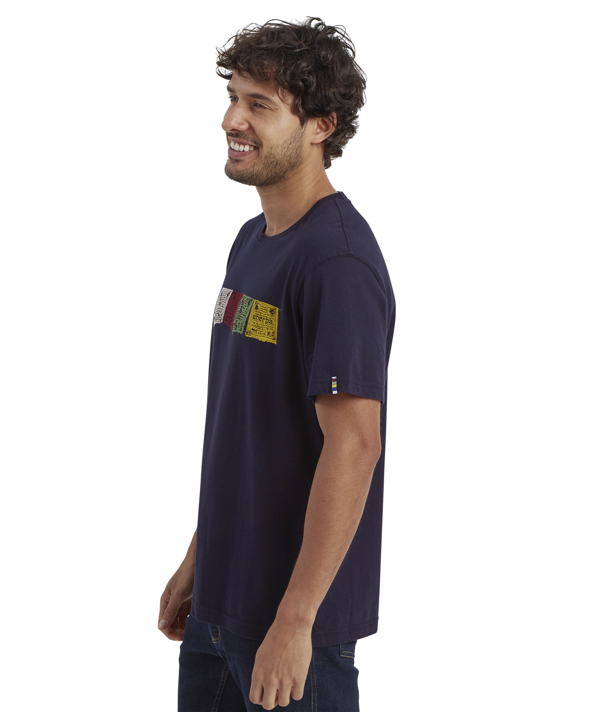 A side-view shot of the Sherpa Adventure Gear Tarcho T-Shirt in Blue, showing its straight hem and clean sleeve cut. The model smiles, with the colorful prayer flag print visible from the side, paired with dark denim for a casual, everyday look.