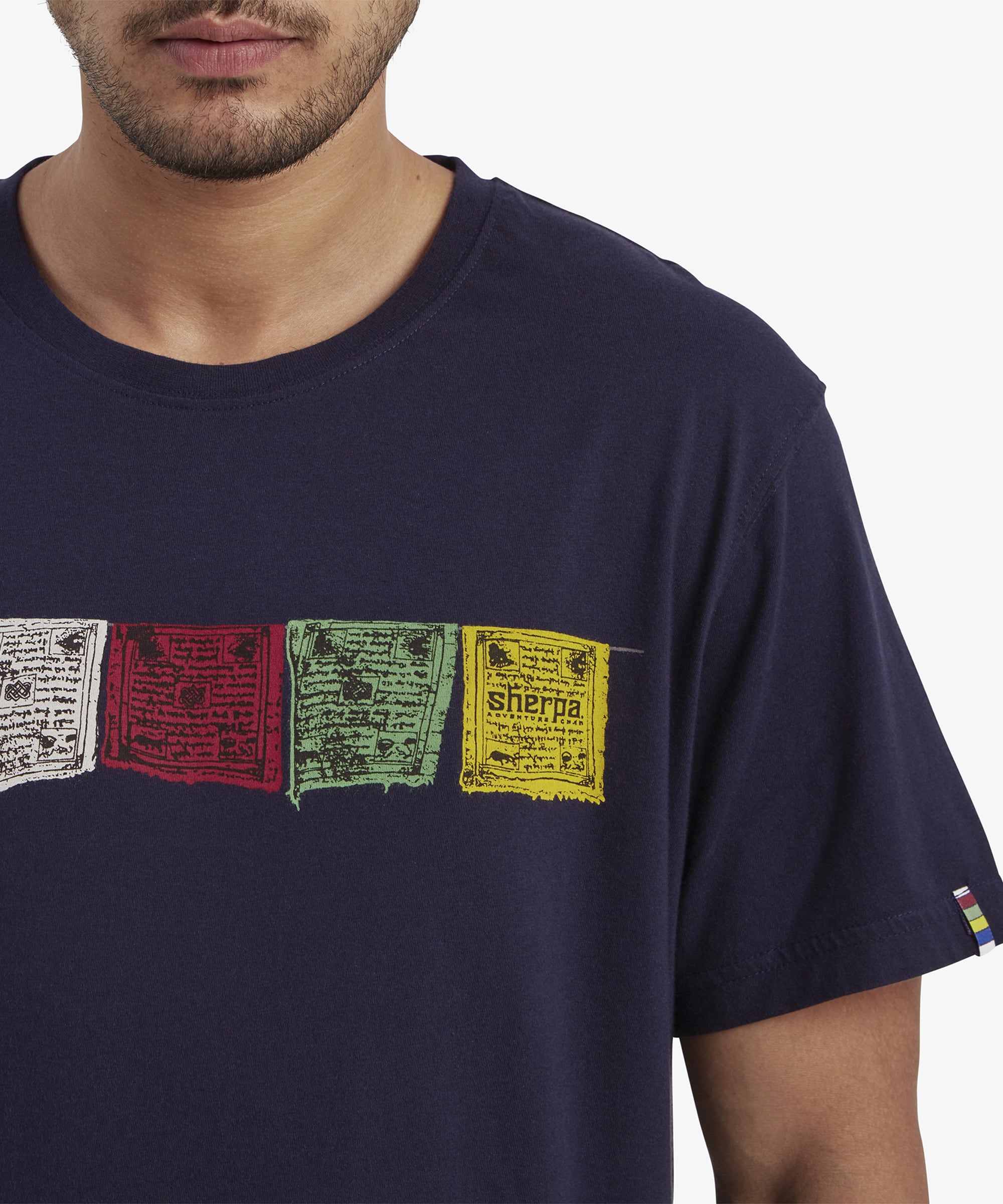 A close-up of the intricate prayer flag designs on the Sherpa Adventure Gear Tarcho T-Shirt in Blue. Each flag showcases detailed script and vibrant colors, emphasising the craftsmanship and cultural significance of the design. A small striped logo detail is visible on the sleeve edge.