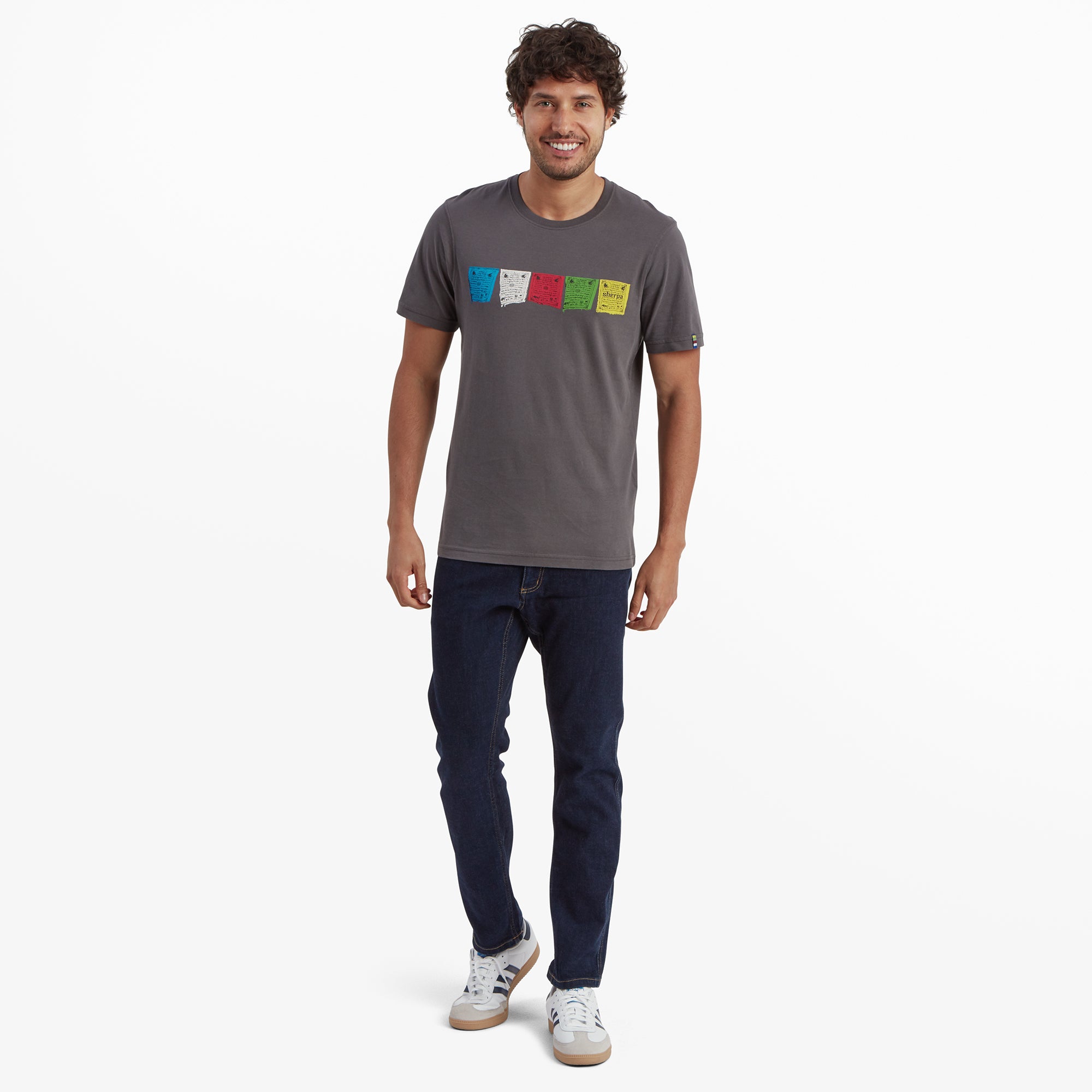 The same male model wearing the Sherpa Adventure Gear Tarcho T-Shirt in Grey, paired with dark blue jeans and white trainers. He is smiling and walking casually, displaying the T-shirt in a relaxed, everyday style.