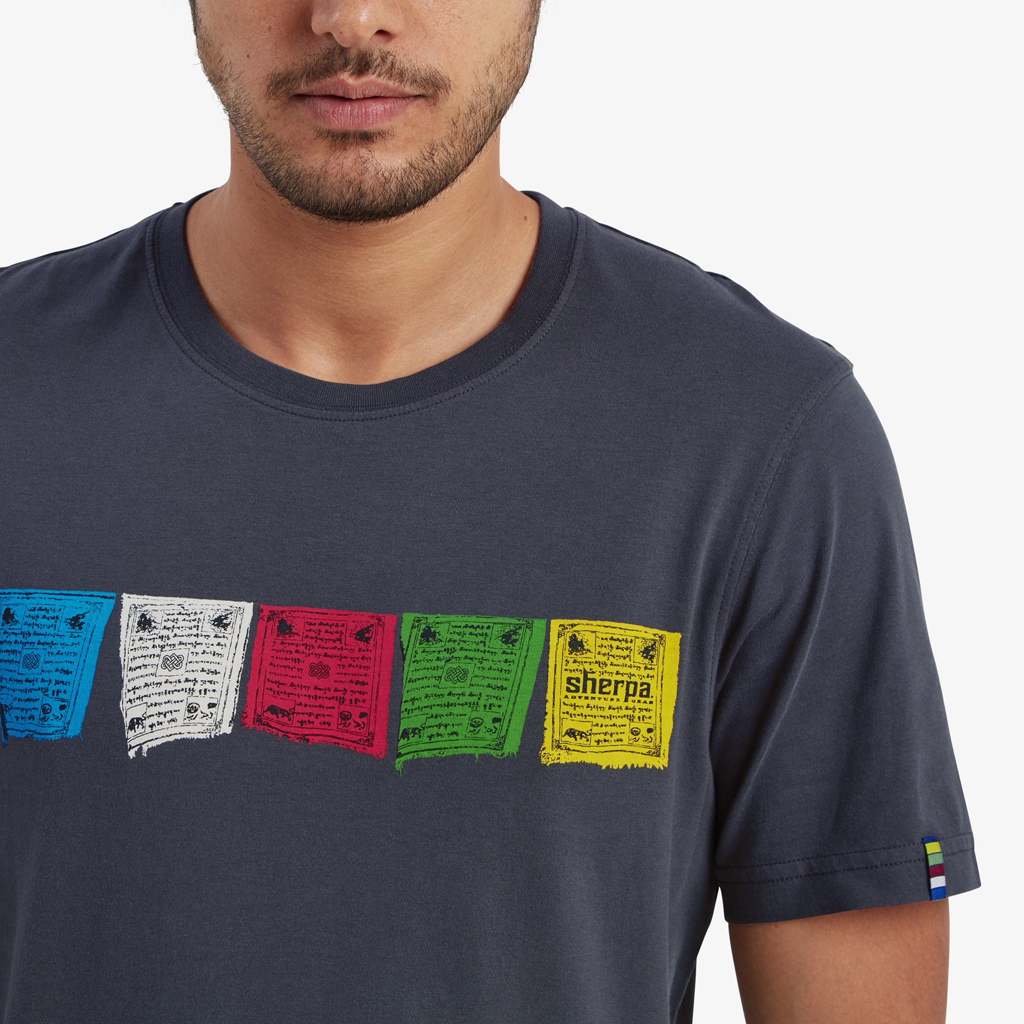 A detailed shot of the colorful Tibetan prayer flags graphic on the Sherpa Adventure Gear Tarcho T-Shirt in Grey. The flags are arranged in a horizontal row with intricate text and symbols, representing Sherpa Adventure Gear's homage to Nepalese heritage.