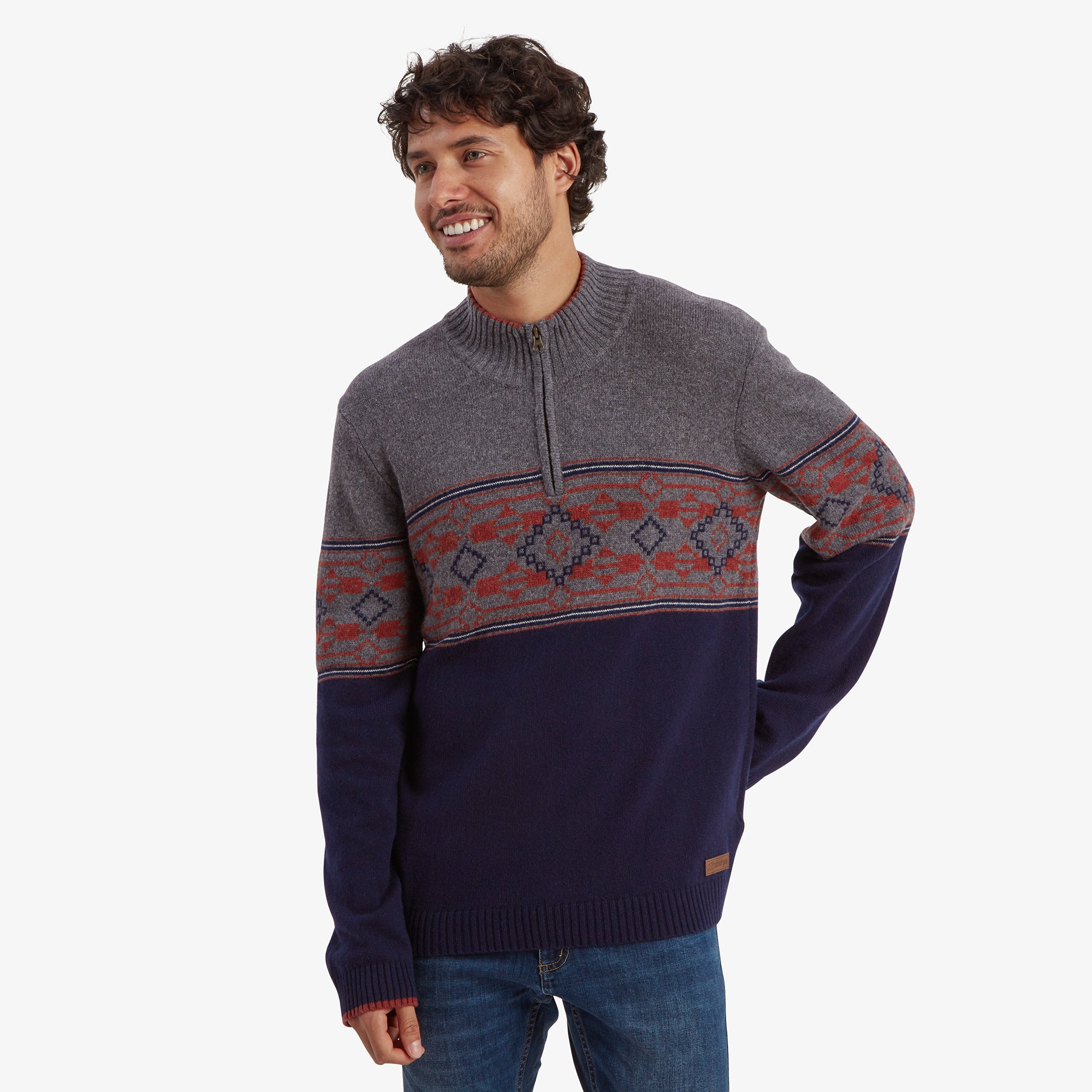 A smiling male model wearing a Sherpa Adventure Gear Tej Eco Quarter Zip Jumper in Blue in a blend of grey and navy blue. The jumper features a patterned band of geometric red, navy, and grey designs across the chest. The collar is half-zip with a ribbed texture, complementing the ribbed cuffs and hem. He pairs the sweater with slim-fit blue jeans, standing confidently.