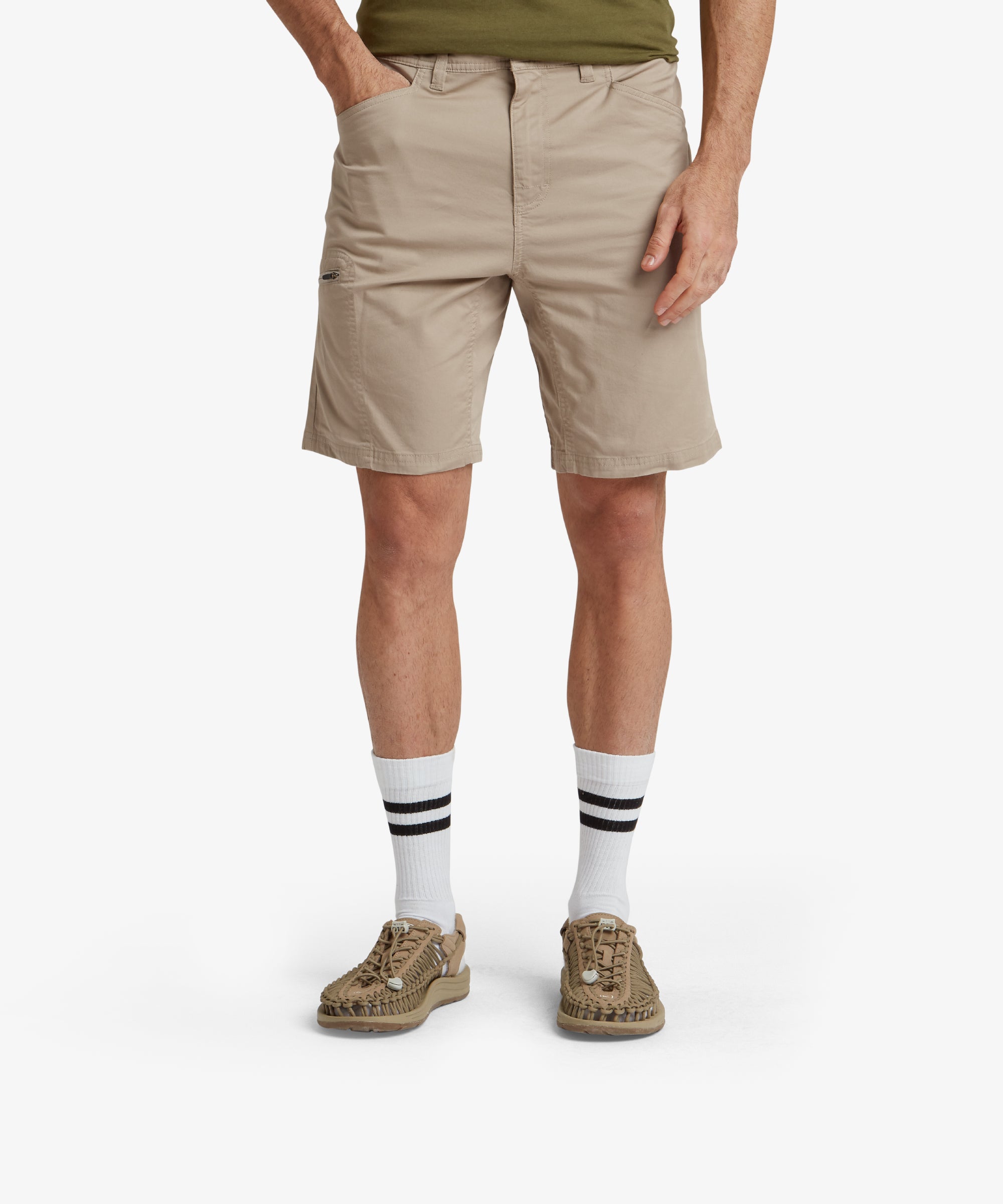 A male model wearing Sherpa Adventure Gear Tenjing Shorts in Beige paired with tan flip-flops. The shorts feature a clean, minimalistic design with front pockets and a single button closure at the waistband. The model is standing upright, showcasing the shorts' length, which ends just above the knee.