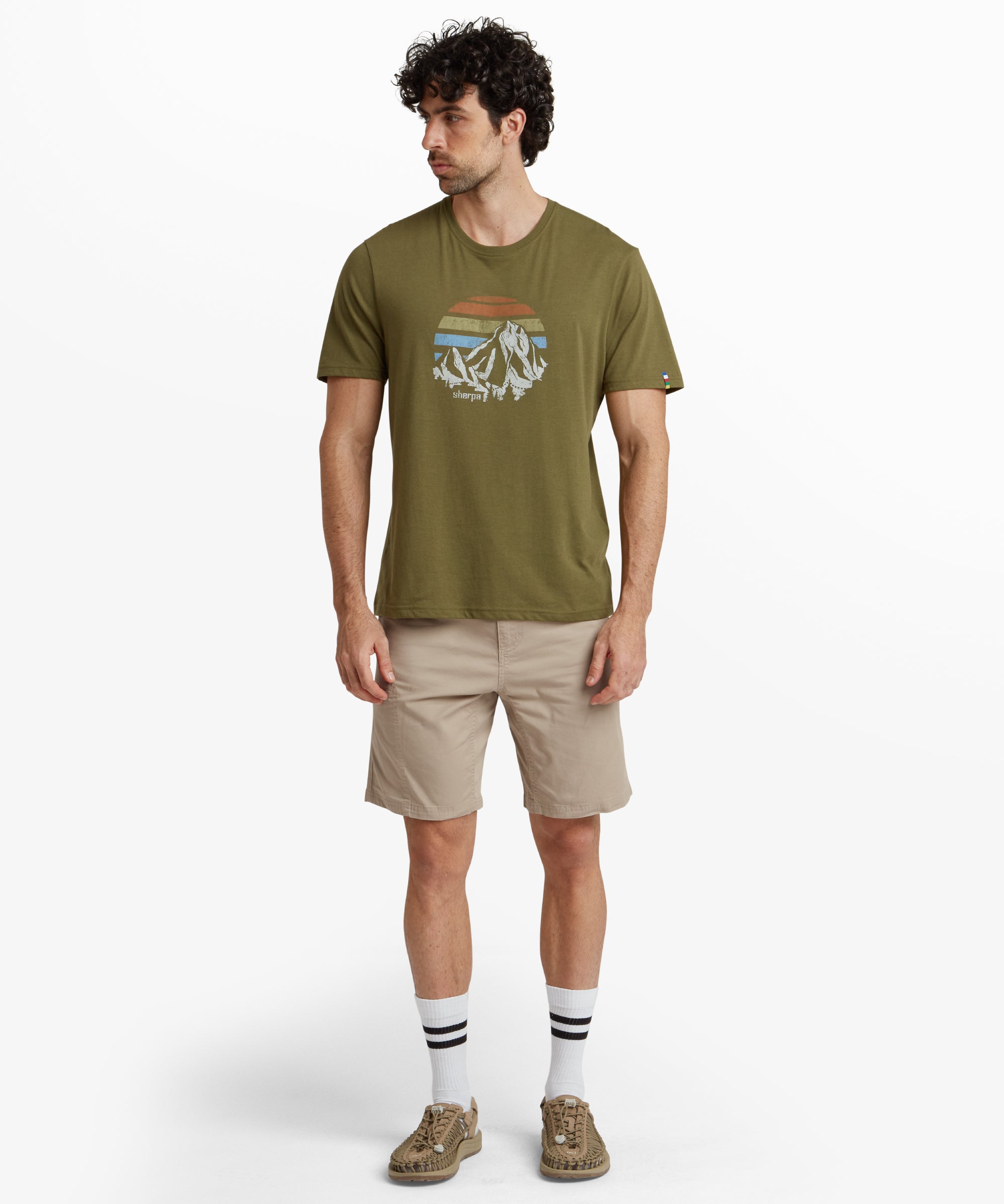 A profile view of the male model wearing Sherpa Adventure Gear Tenjing Shorts in Beige, emphasising the tailored fit and side zipper pocket detail on the thigh. The back pocket is slightly visible, and the model's flip-flops complement the casual look.