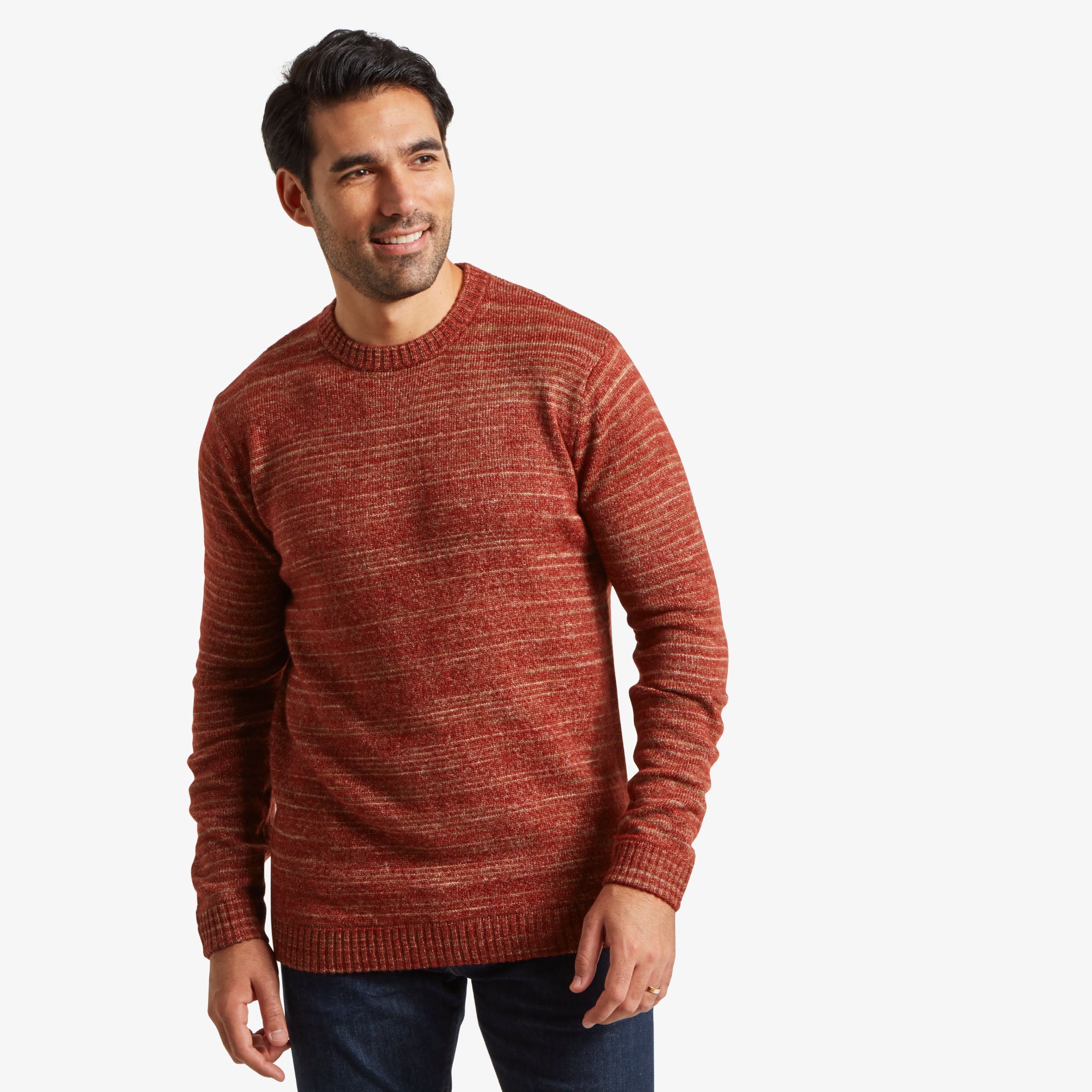 A man with short dark hair, neatly styled, is wearing a Sherpa Adventure Gear Vidvan Crew in Brown. He is standing against a plain white background, smiling slightly and looking off to the side. The sweater fits comfortably and showcases a textured knit with horizontal marled stripes. His posture is relaxed, with one arm at his side and the other slightly bent. He pairs the sweater with dark jeans.