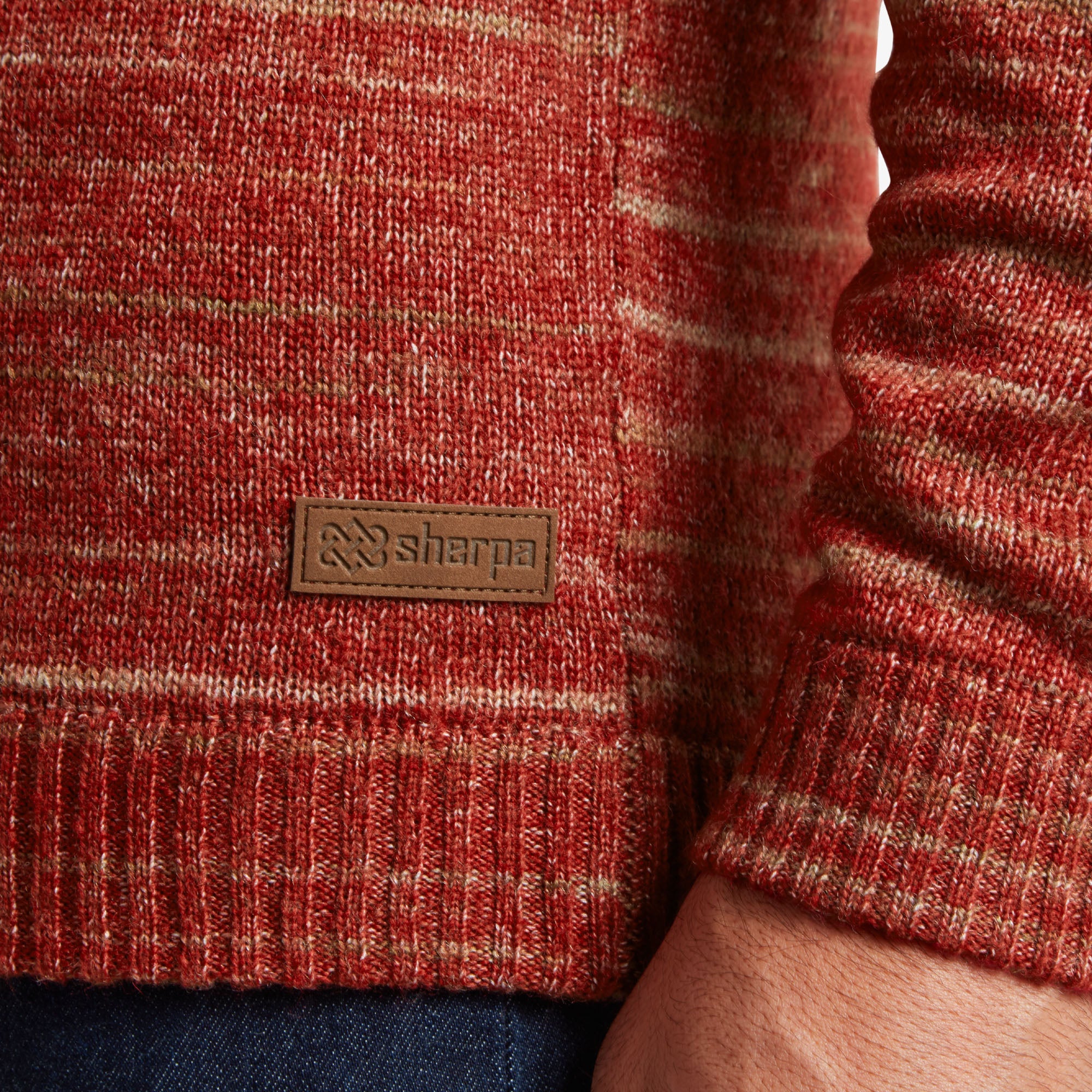 A close-up of the bottom hem of the Sherpa Adventure Gear Vidvan Crew in Brown, highlighting the Sherpa Adventure Gear brand tag. The small, rectangular, brown leather patch is stitched to the ribbed hem and embossed with the Sherpa logo. The marled knit design continues in the background, with the texture of the knit clearly visible. Part of the man’s hand and arm is seen, along with the ribbed cuff of the sweater's sleeve.