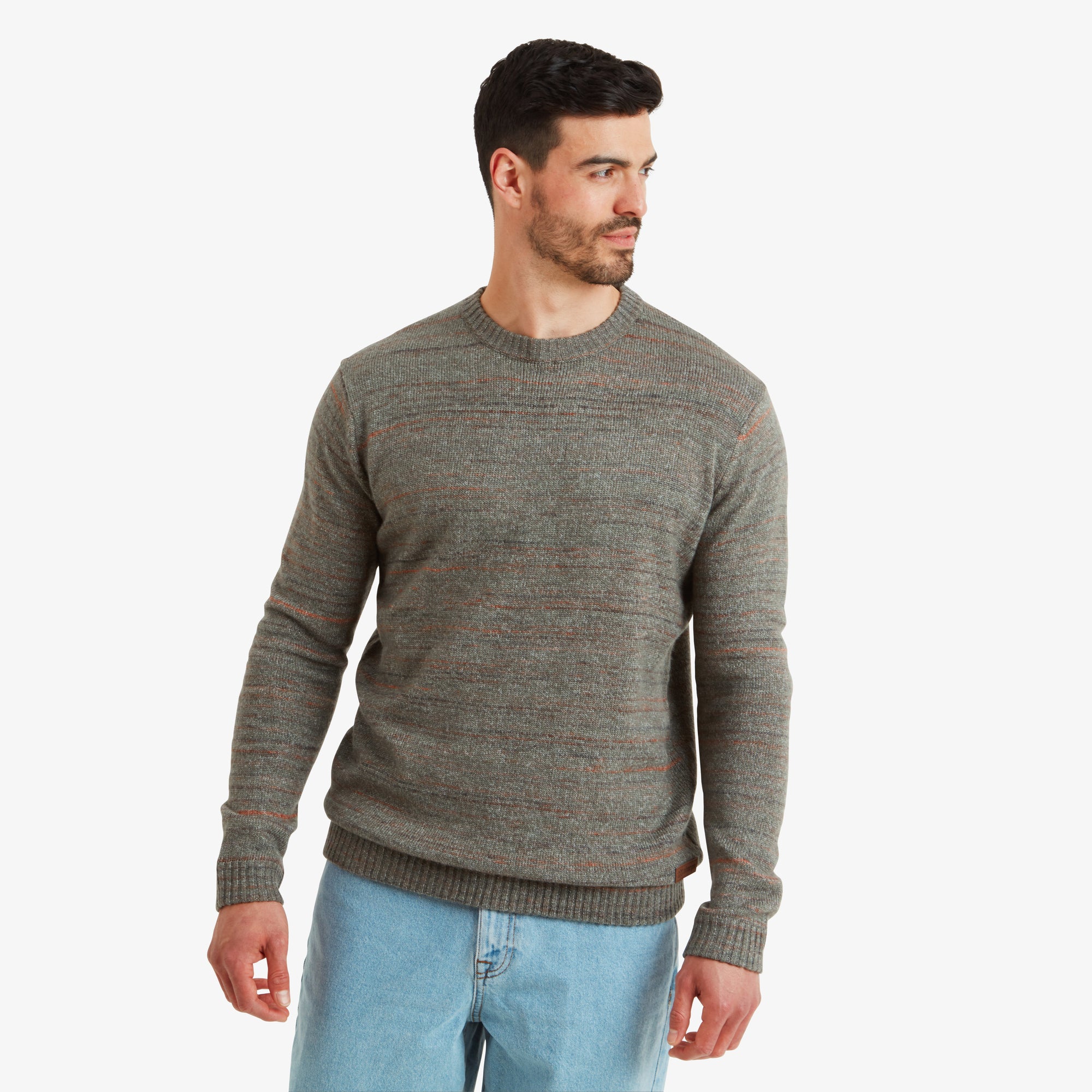 A front-facing view of a man wearing a Sherpa Adventure Gear Vidvan Crew in Grey with subtle orange thread accents. The sweater has a fine knit texture and a fitted silhouette, complemented by light blue denim jeans. The man gazes confidently to the side with a slight smile.