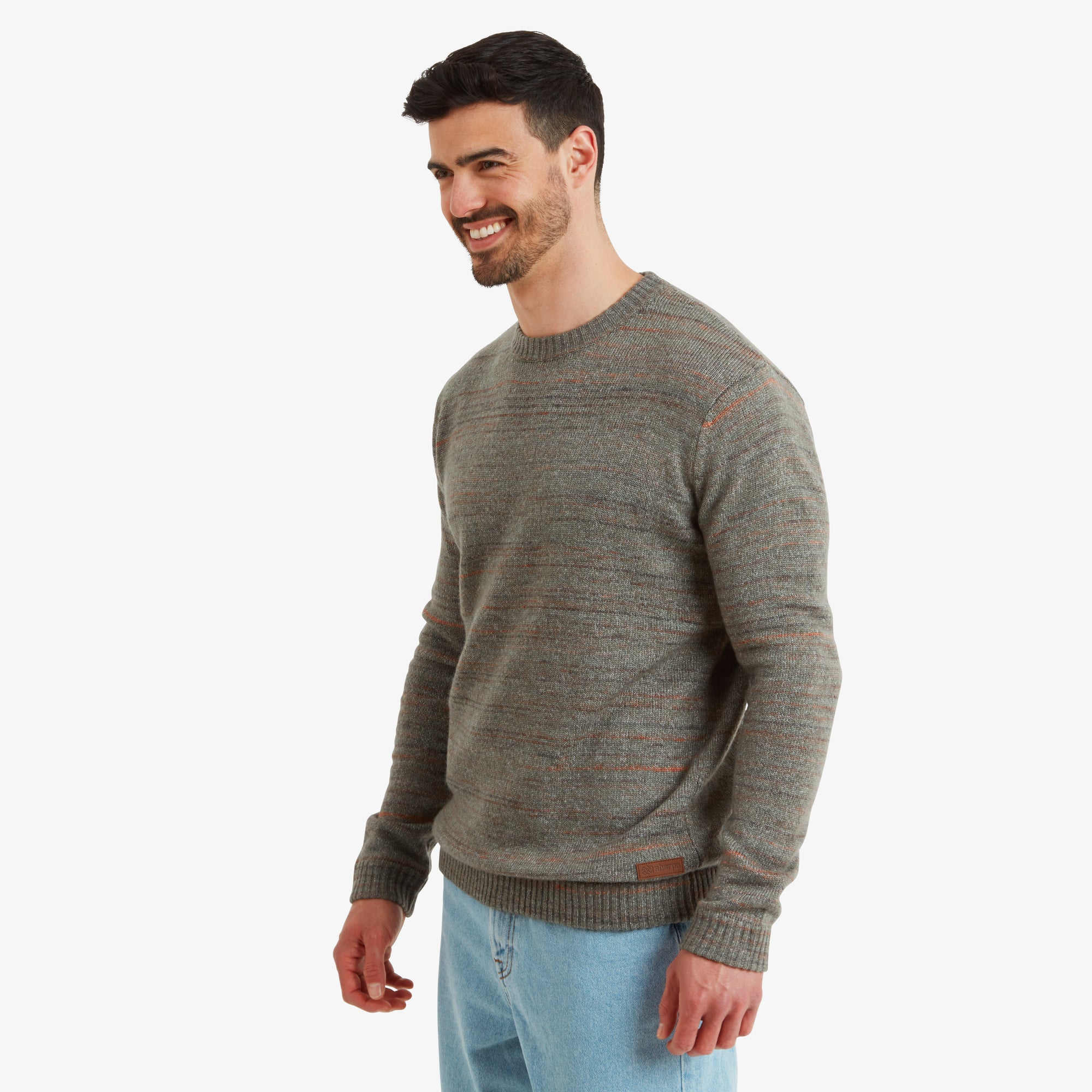 A side profile view of the man wearing the Sherpa Adventure Gear Vidvan Crew in Grey, showcasing its clean side seams and fitted cuffs. The orange accents and knit detailing are subtly visible. The man smiles warmly while looking ahead.