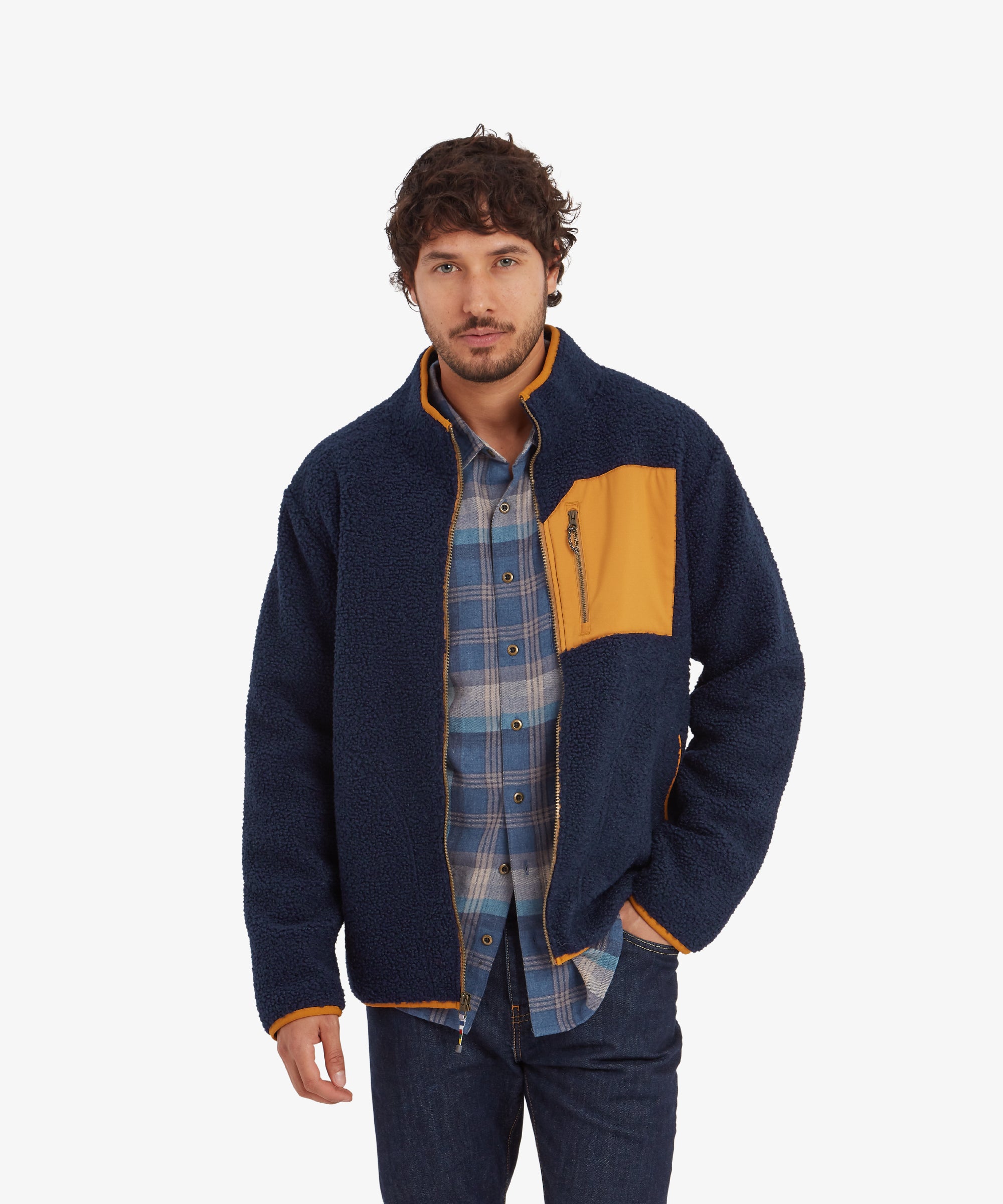 A man modeling a Sherpa Adventure Gear Yatri Full Zip Jacket in Blue with a mustard yellow chest pocket and zip. The jacket has contrasting orange piping along the edges. He is wearing a blue plaid shirt underneath, paired with dark denim jeans. He stands with a neutral expression, hands in the side pockets.