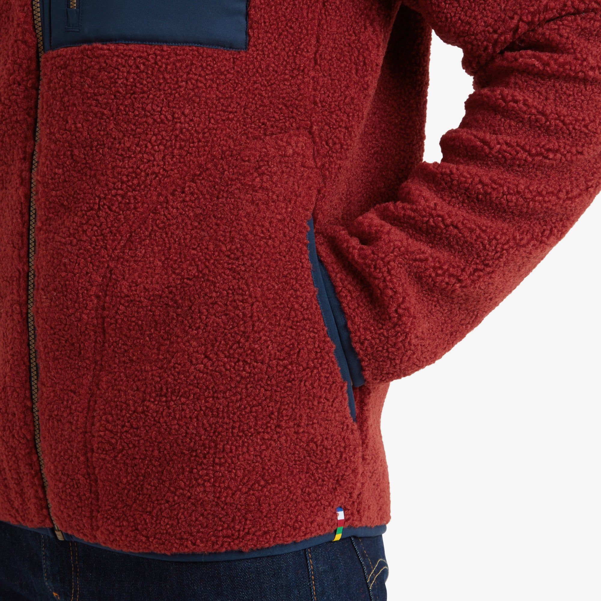 A detailed view of the fleece material around the hand pocket on the Sherpa Adventure Gear Yatri Full Zip Jacket in Light Brown, with navy trim and multicolored Sherpa-inspired stitching near the hem.