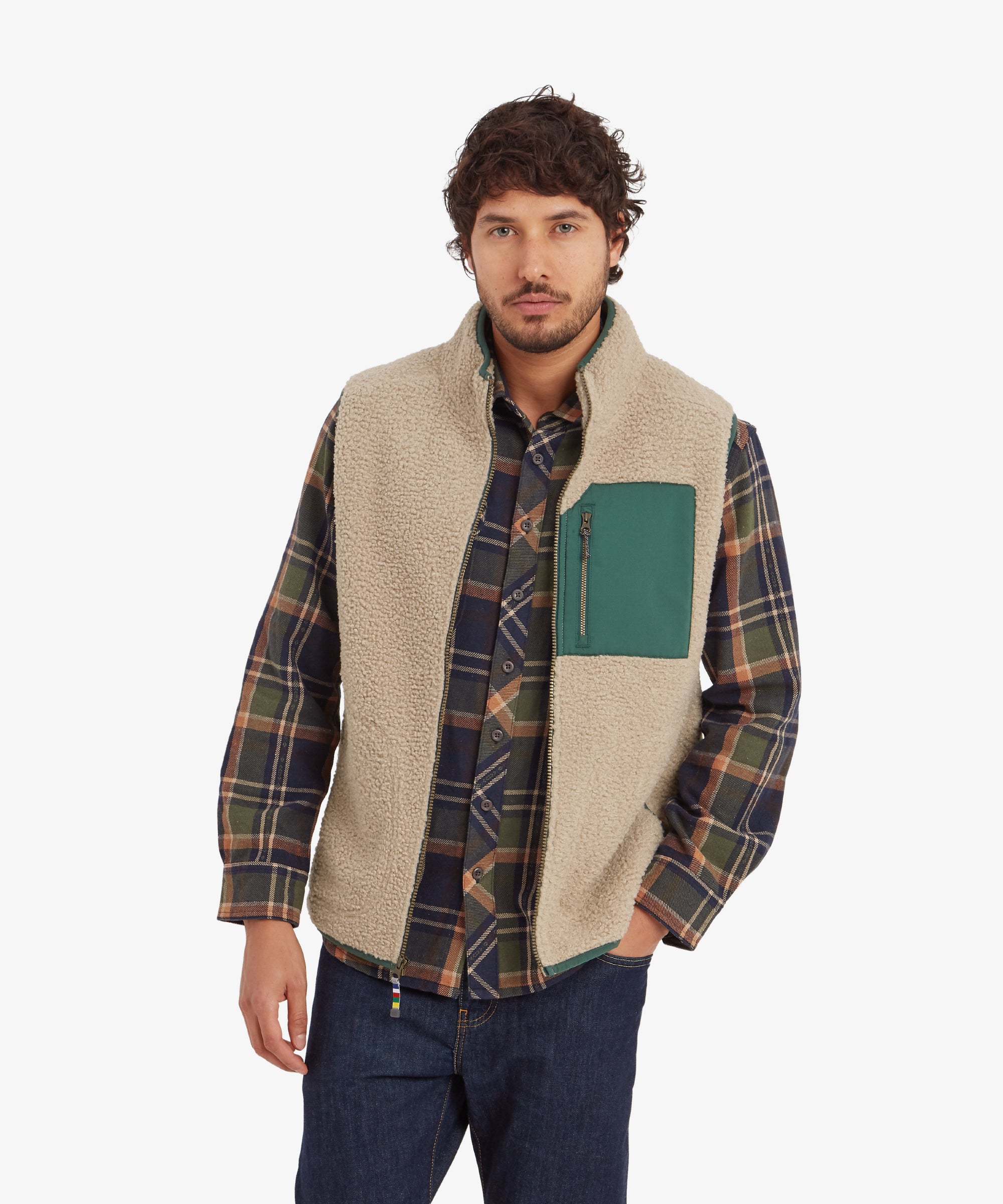 A man wearing a Sherpa Adventure Gear Yatri Vest in Beige with a forest green chest pocket. The fleece texture is soft and fluffy, with the green chest pocket featuring a zipper for storage. He pairs it with a plaid shirt in earthy tones of green and brown, adding a casual outdoor vibe. His left hand is resting in the side pocket of the gilet.