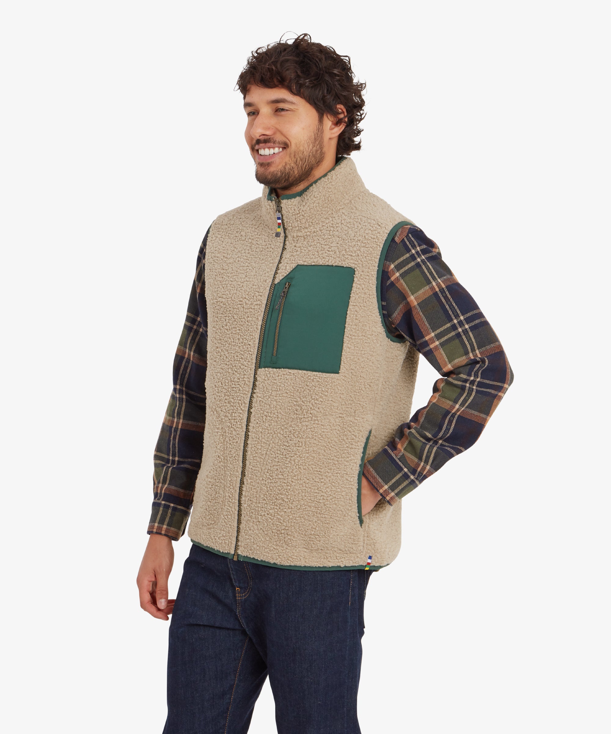 A three-quarter profile view of the man, showcasing the green chest pocket of the Sherpa Adventure Gear Yatri Vest in Beige prominently. He is smiling and has his left hand casually placed in the side pocket of the gilet. The plaid shirt sleeves provide contrast against the beige fleece texture.