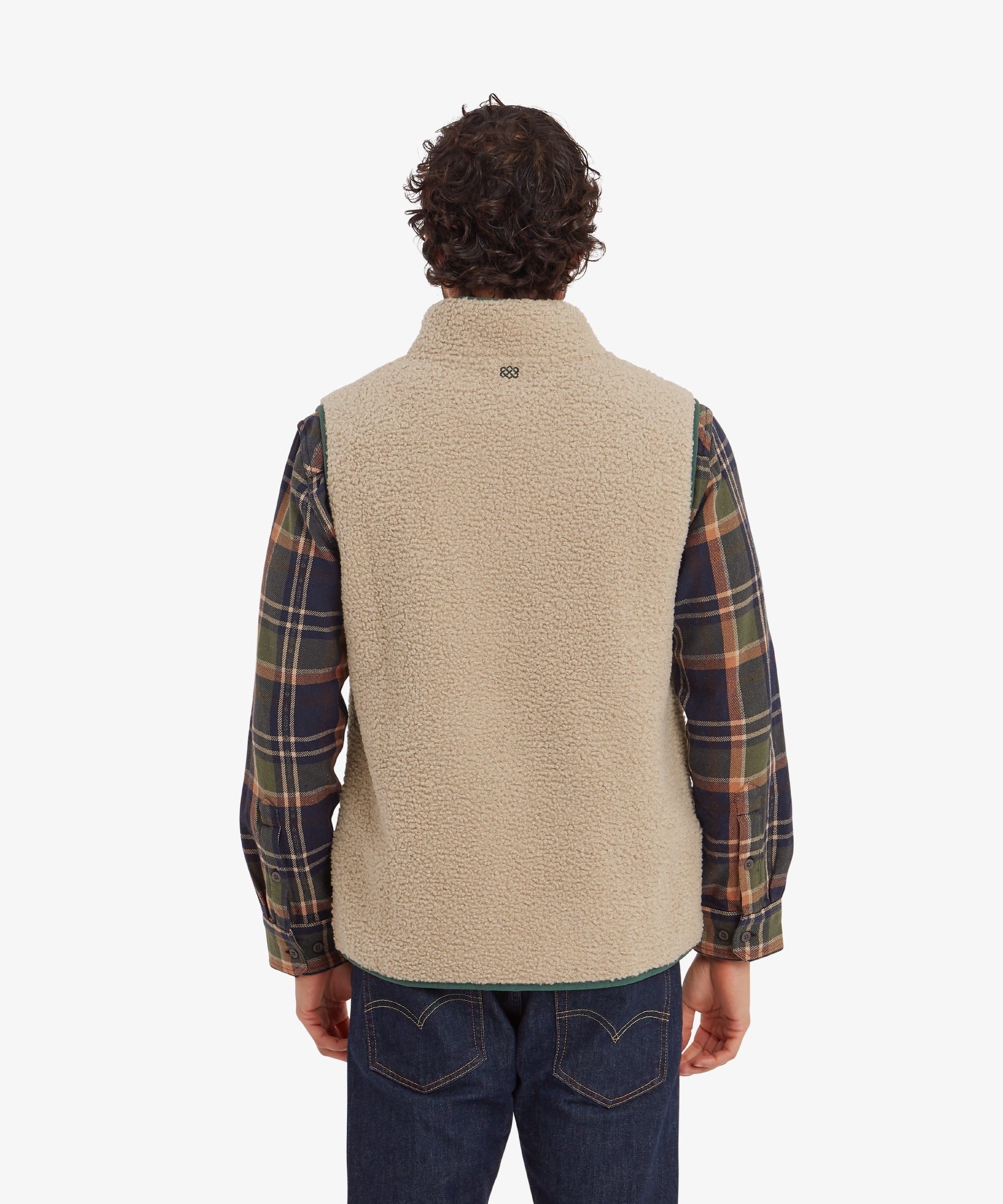 A rear view of the Sherpa Adventure Gear Yatri Vest in Beige, highlighting its clean and simple back design. The fleece extends smoothly, with a subtle logo detail at the nape. The man is wearing the same plaid shirt underneath, which adds color and pattern.