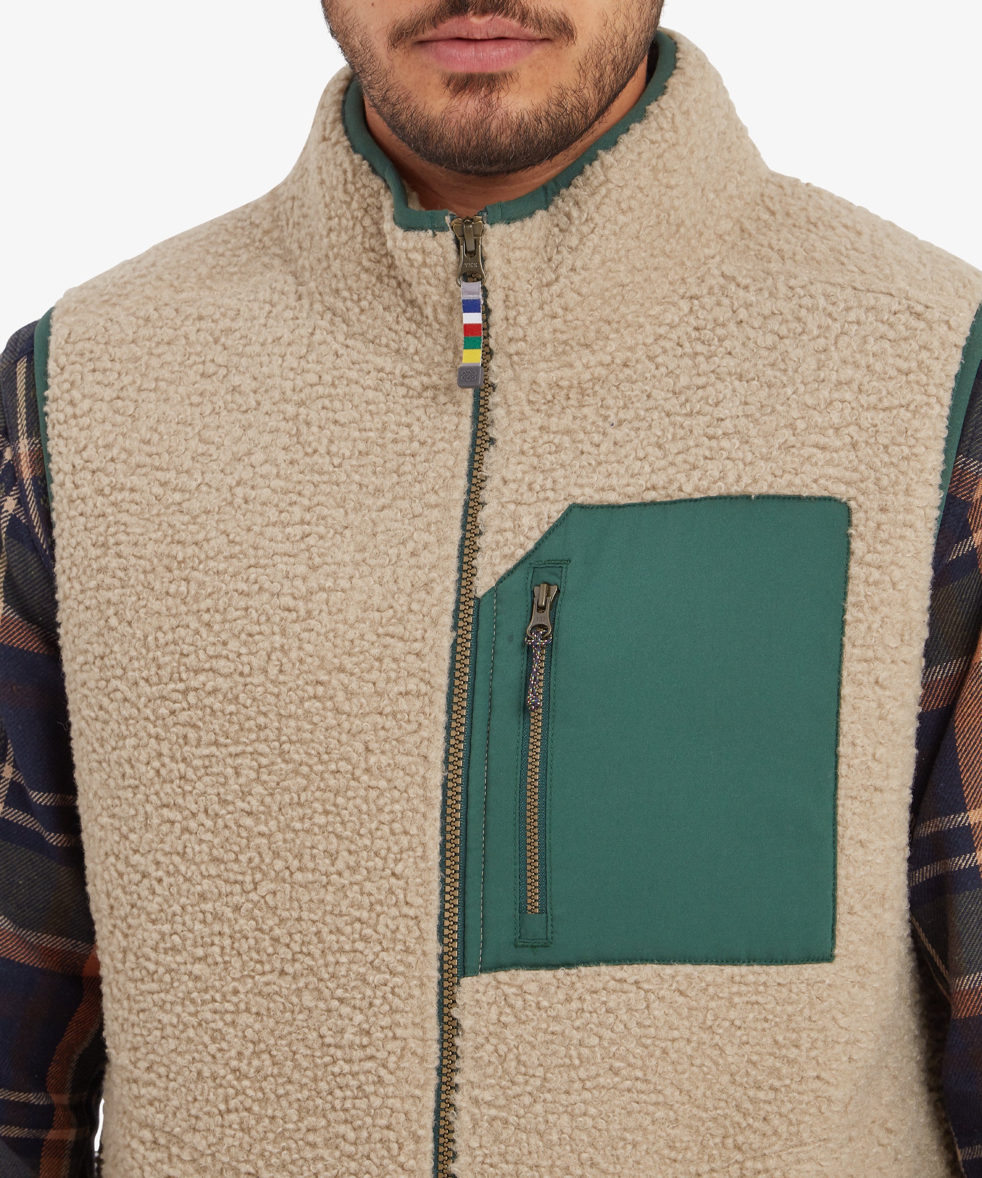 A close-up of the chest area, focusing on the texture of the Sherpa Adventure Gear Yatri Vest in Beige and the green chest pocket with a zipper. The multicolored striped zipper pull adds a playful touch to the functional design.