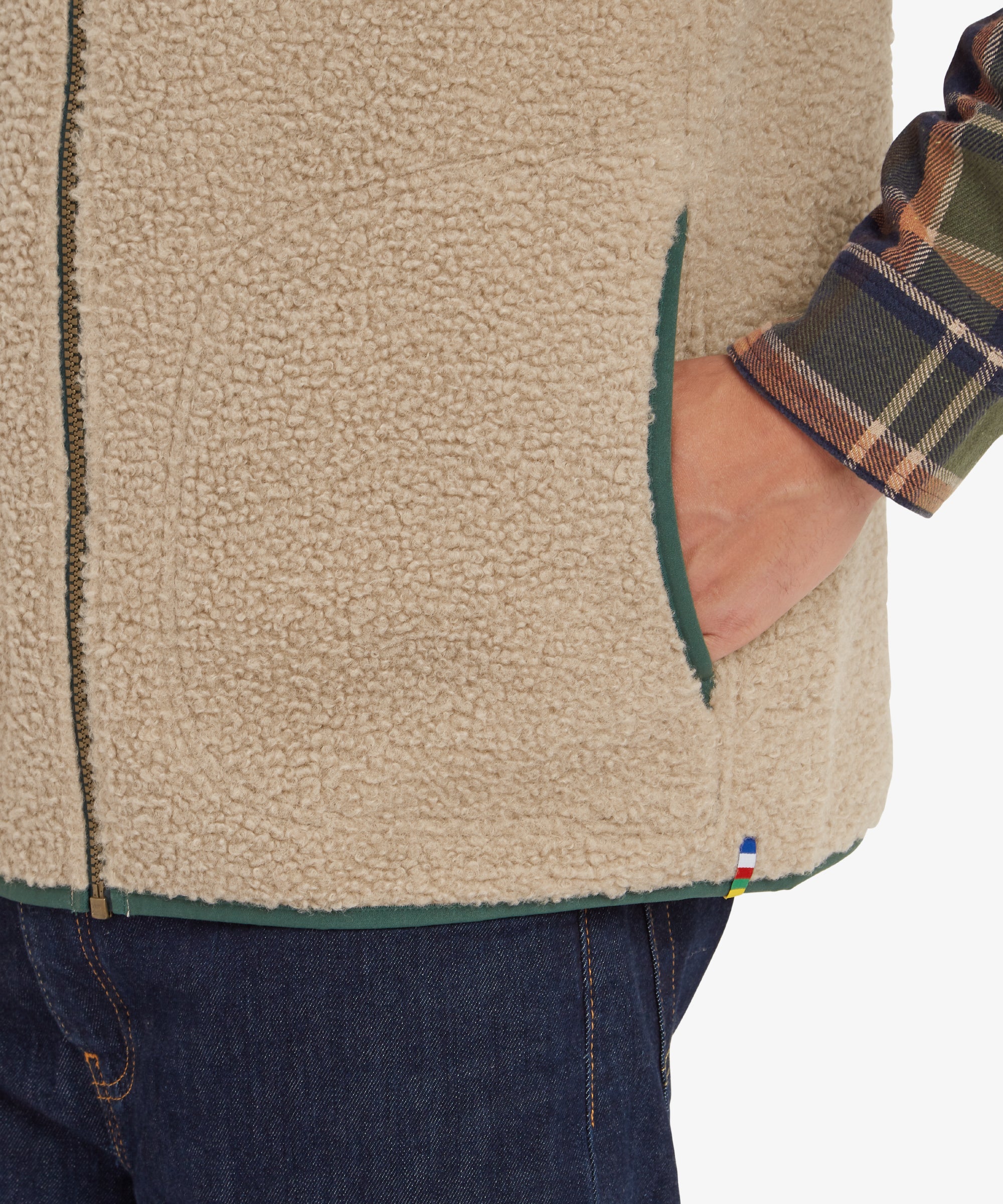 A detailed view of the lower portion of the Sherpa Adventure Gear Yatri Vest in Beige, emphasising the side pocket where the man’s hand is resting. The edges of the fleece are trimmed with green fabric, matching the chest pocket, and a small multi-colored flag tag is visible on the hem.