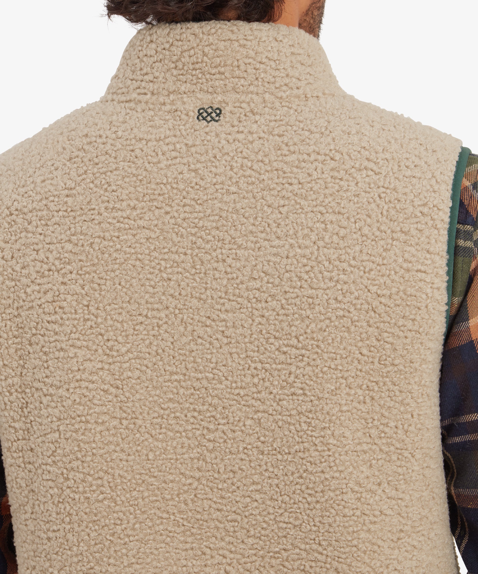 A zoomed-in view of the back upper portion of the Sherpa Adventure Gear Yatri Vest in Beige, showcasing the Sherpa texture and a small embroidered logo at the center near the neckline. The plaid shirt collar adds a subtle, contrasting pattern to the soft beige fleece.