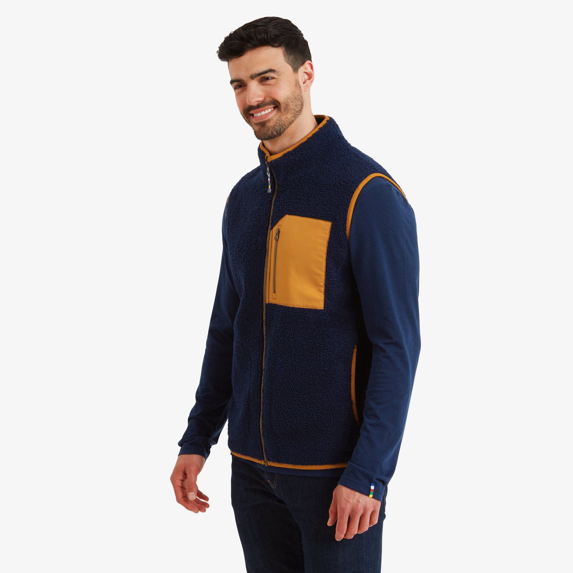 Side profile of the man in the Sherpa Adventure Gear Yatri Vest in Blue. The vest’s slim fit and mustard yellow side detailing are highlighted. The man has his hands in his pockets and is smiling slightly, showcasing the functional yet stylish design.