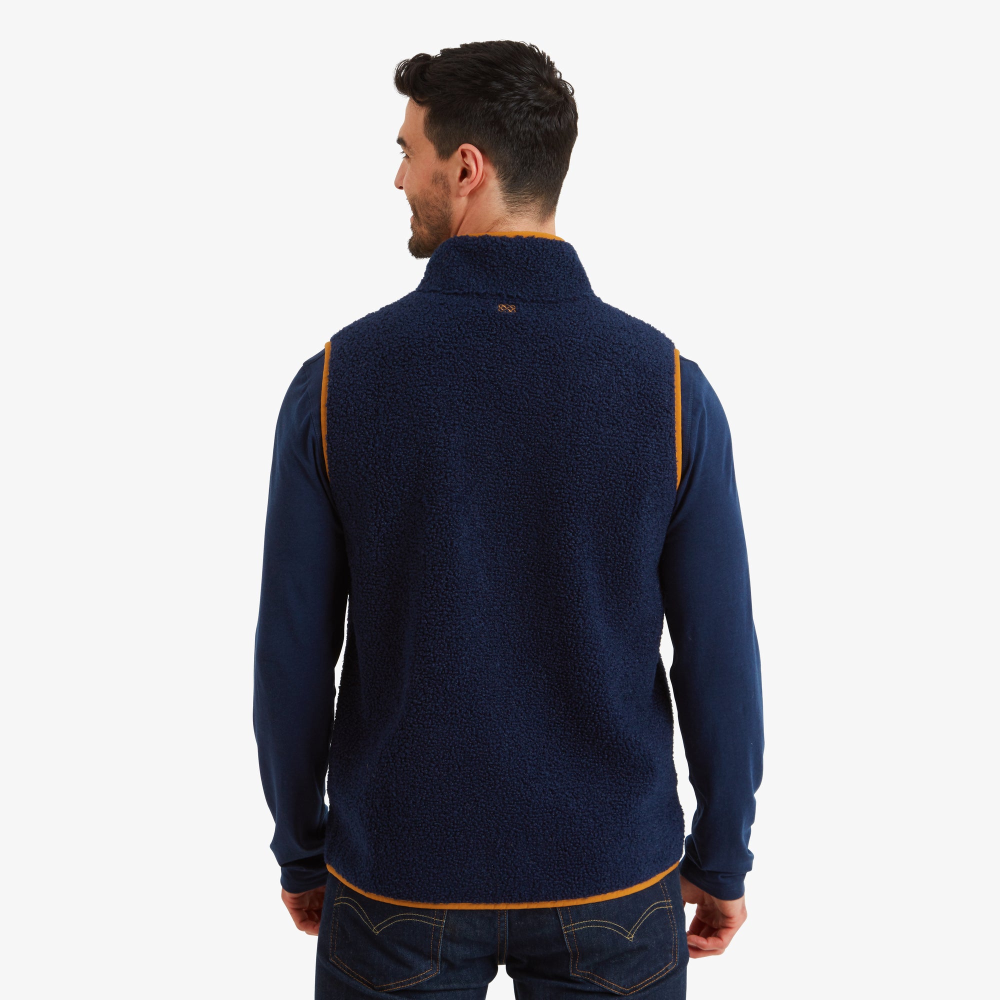 Rear view of the Sherpa Adventure Gear Yatri Vest in Blue. The back of the vest is entirely navy blue, with a small embroidered Sherpa logo near the collar and orange piping around the edges. The vest pairs seamlessly with the navy long-sleeve shirt and jeans.