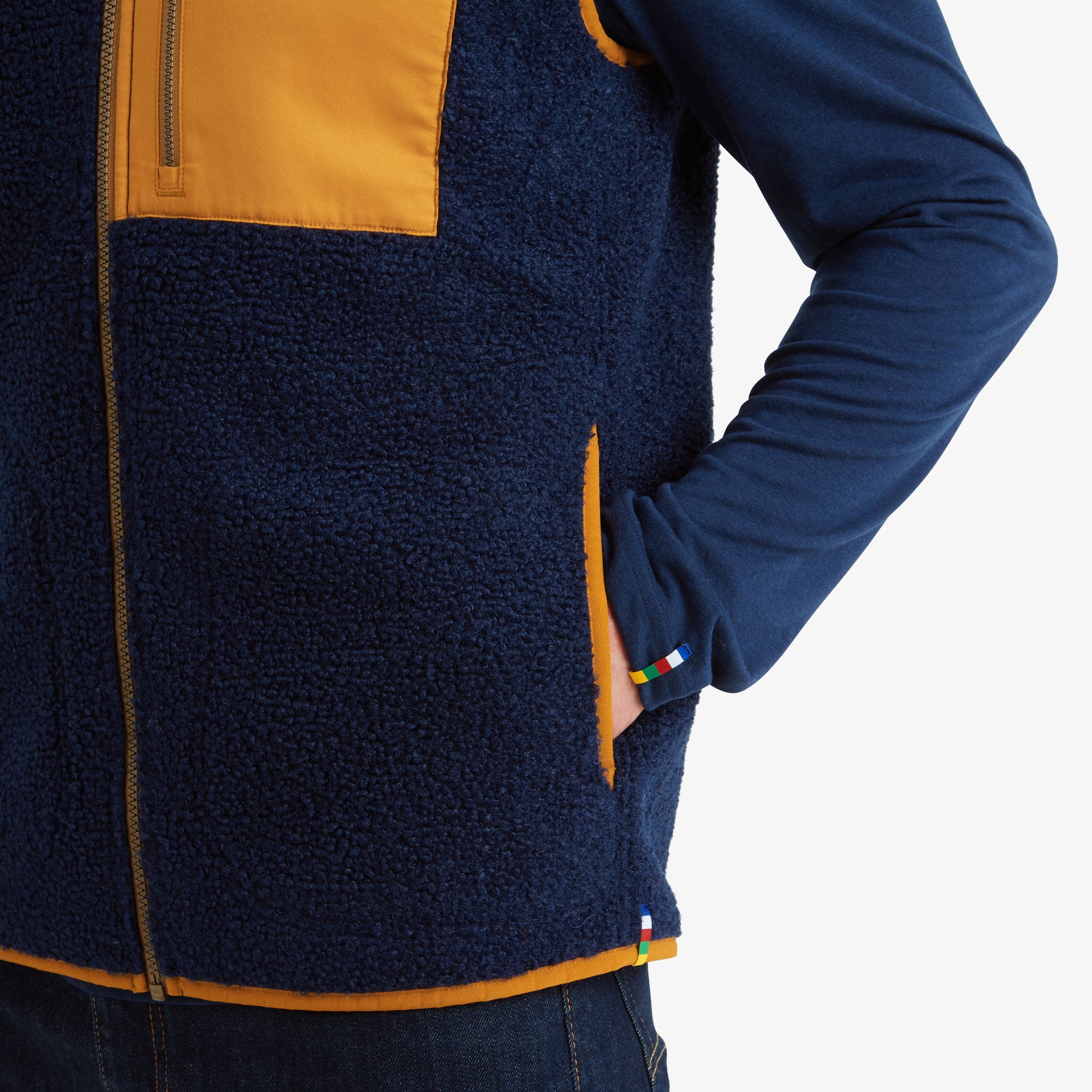 Close-up of the hand pocket with colorful accent stitching. The man has his hand in the side pocket of the Sherpa Adventure Gear Yatri Vest in Blue, revealing a small multicolored stripe detail near the hem. The vest’s orange piping and plush fabric texture are also visible in the shot.