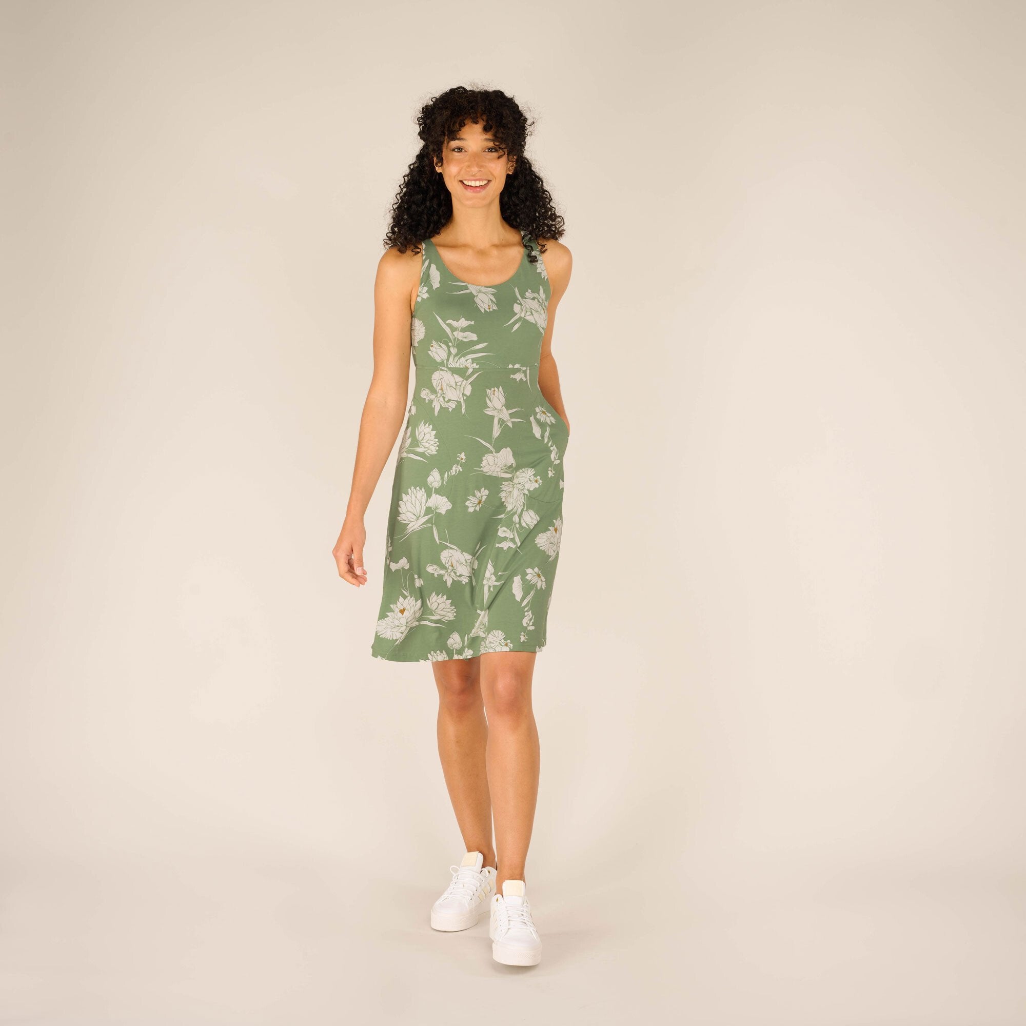 A woman with curly black hair smiles while walking forward, wearing a Sherpa Adventure Gear Neha Dress in Green with a white floral pattern. The dress has a fitted bodice and flares slightly at the bottom. She pairs it with white sneakers and poses against a neutral beige background.