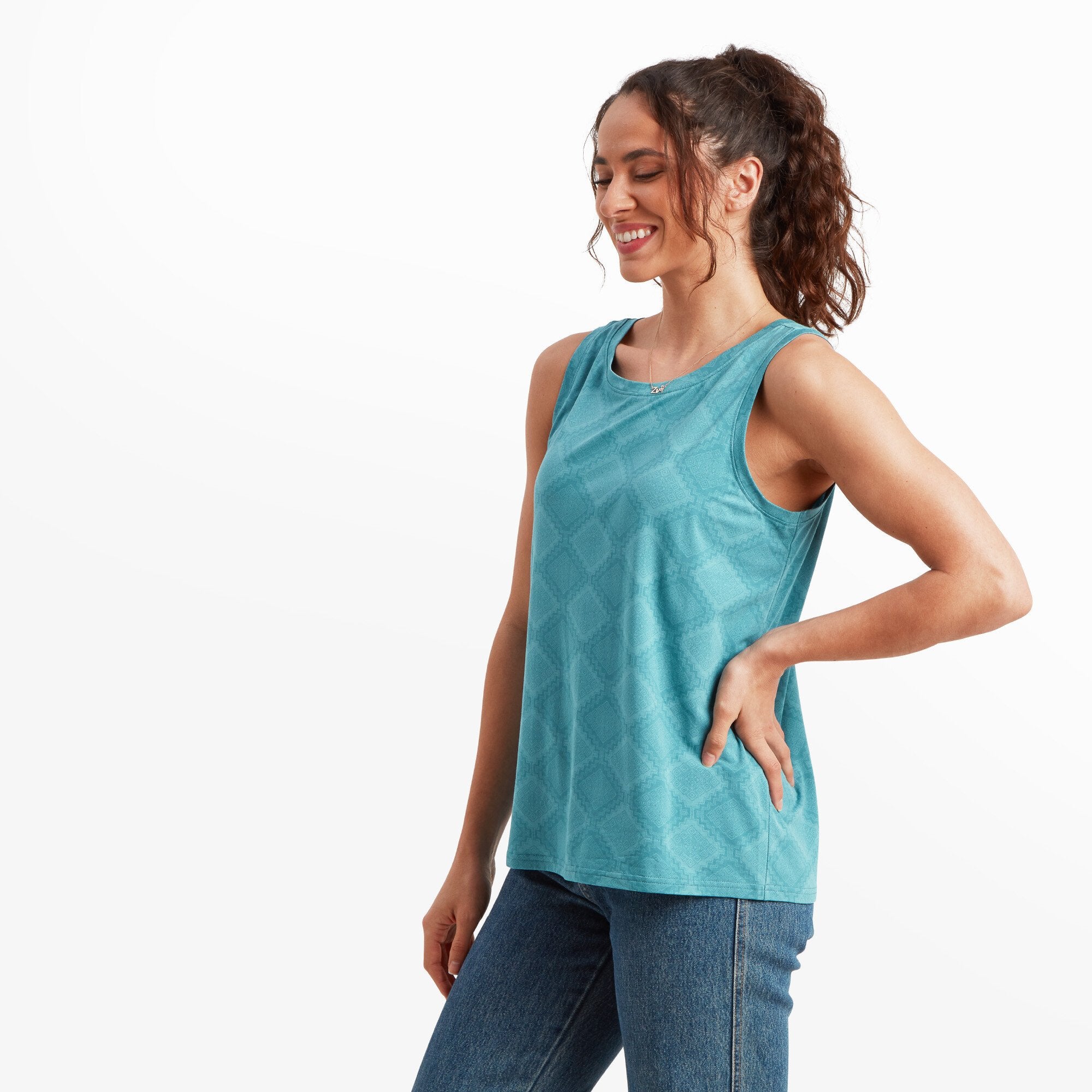 The same woman is shown from the side, slightly turned to the left with her hand resting on her hip. The Sherpa Adventure Gear Neha High Neck Tank in Blue highlights the geometric diamond pattern, showcasing its sleeveless cut and slightly loose fit around the torso. Her ponytail adds to the casual and summery feel.