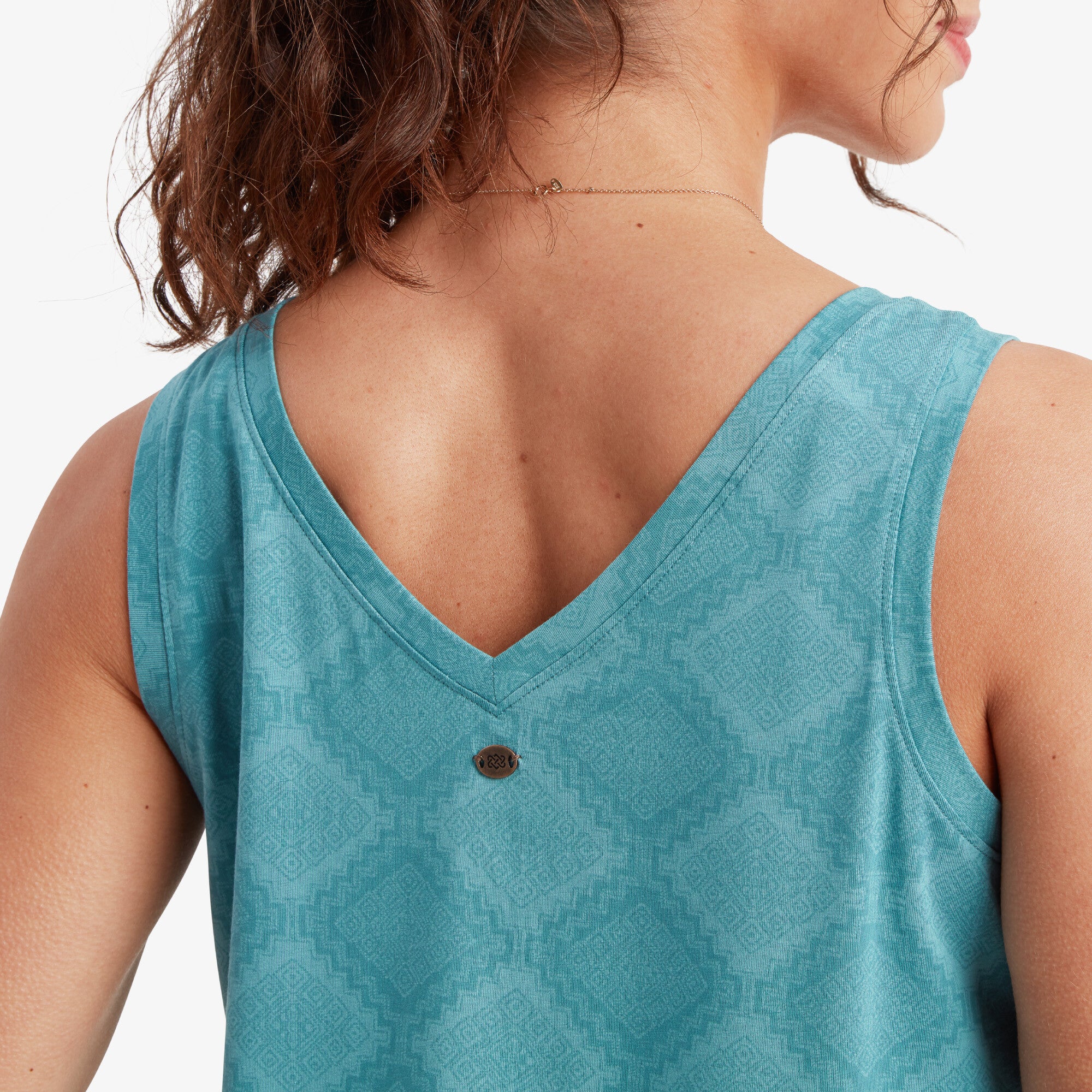 A zoomed-in view of the back of the Sherpa Adventure Gear Neha High Neck Tank in Blue, focusing on the V-shaped neckline and the small metallic logo accent. The intricate diamond-pattern texture of the fabric is clearly visible, emphasising the craftsmanship and design details. The model's delicate necklace and exposed shoulders enhance the focus on the top’s styling.