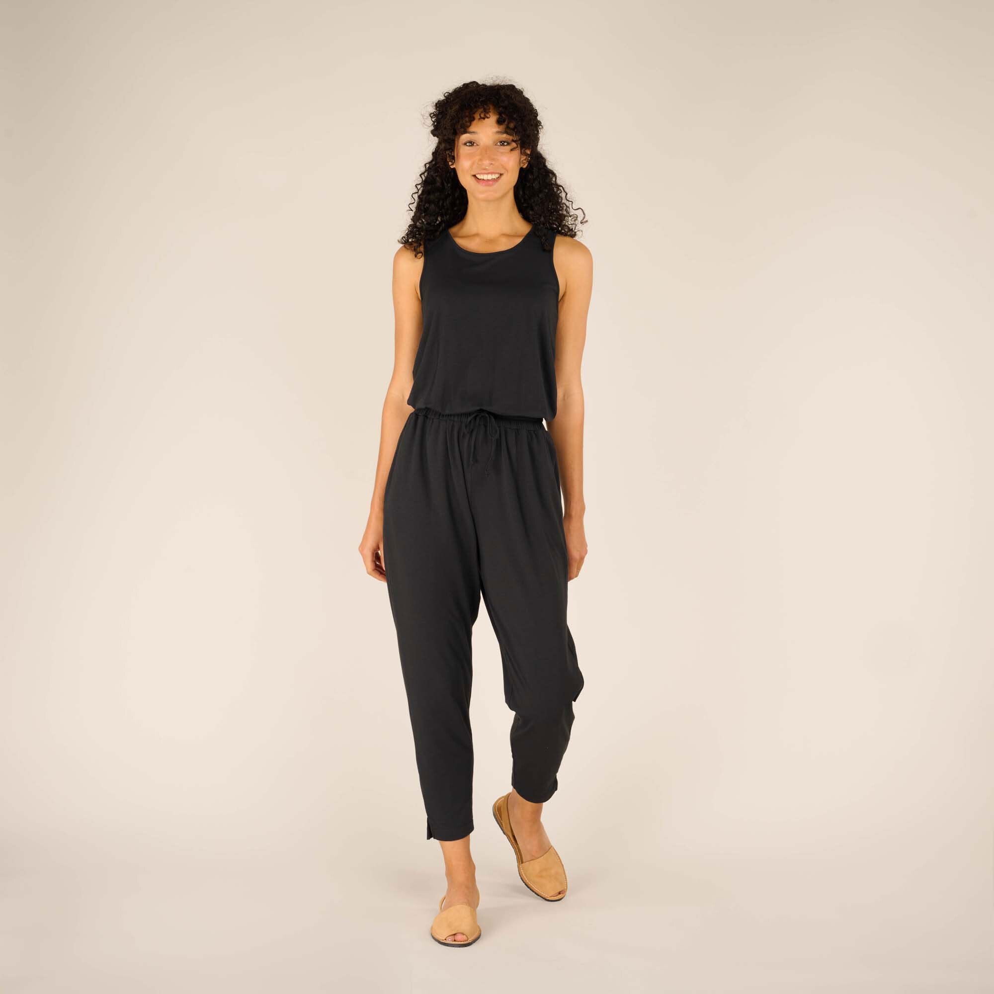 A woman wearing a sleek, Sherpa Adventure Gear Neha Jumpsuit in Black with a relaxed fit. The jumpsuit features a drawstring waistband, tapered legs, and side pockets. She pairs the outfit with tan flat sandals, standing in a neutral studio setting while smiling confidently.