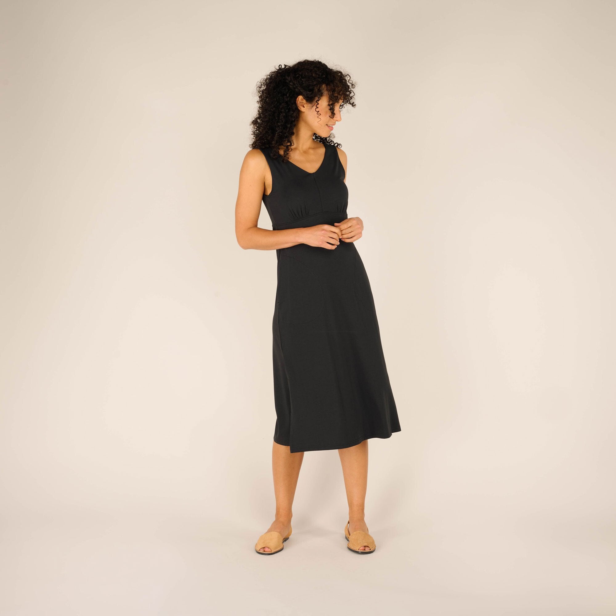 A woman with curly dark hair wears a Sherpa Adventure Gear Neha Midi Dress in Black with a V-neckline and sleeveless design. She stands with her hands gently clasped in front of her, looking slightly to the side. The dress is fitted at the waist and flares slightly, falling just below the knee. She wears tan open-toe sandals.