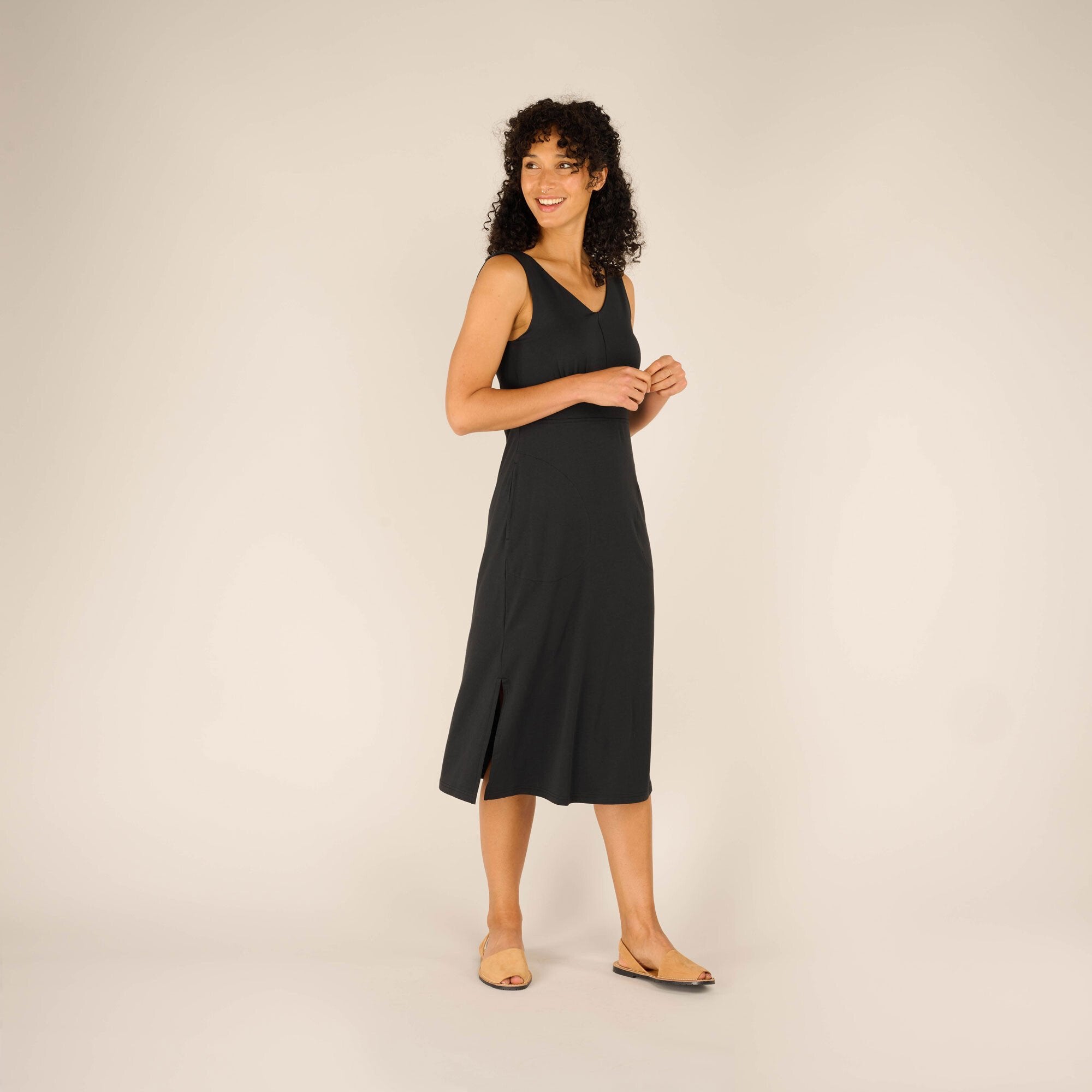 The model is turned slightly to the right, offering a side view of the Sherpa Adventure Gear Neha Midi Dress in Black. She smiles warmly while looking off-camera. The dress has subtle side slits near the hem, adding to its flowy silhouette. She keeps her arms relaxed at her sides.