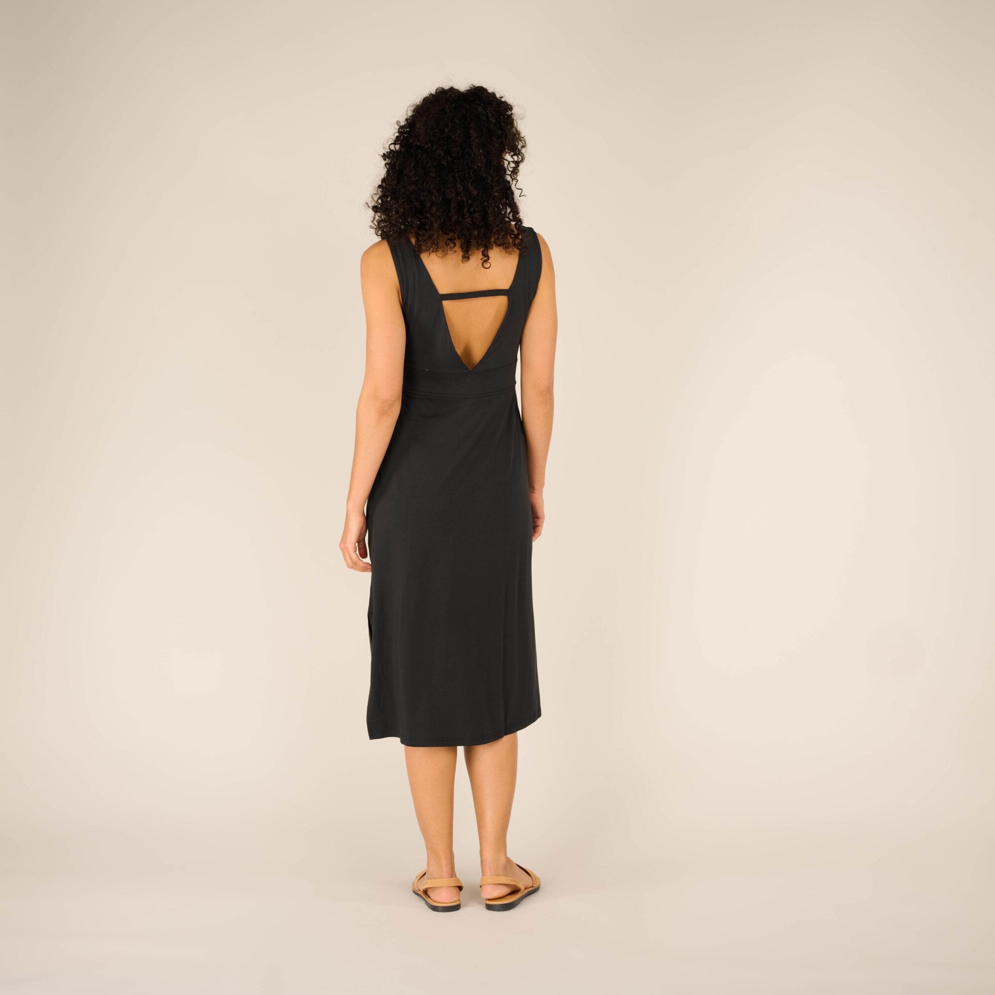 The model is shown from behind, revealing the deep V-back cut of the Sherpa Adventure Gear Neha Midi Dress in Black with a thin horizontal strap across the upper back for added support. The fitted waistline enhances the silhouette, and the hem reaches mid-calf.