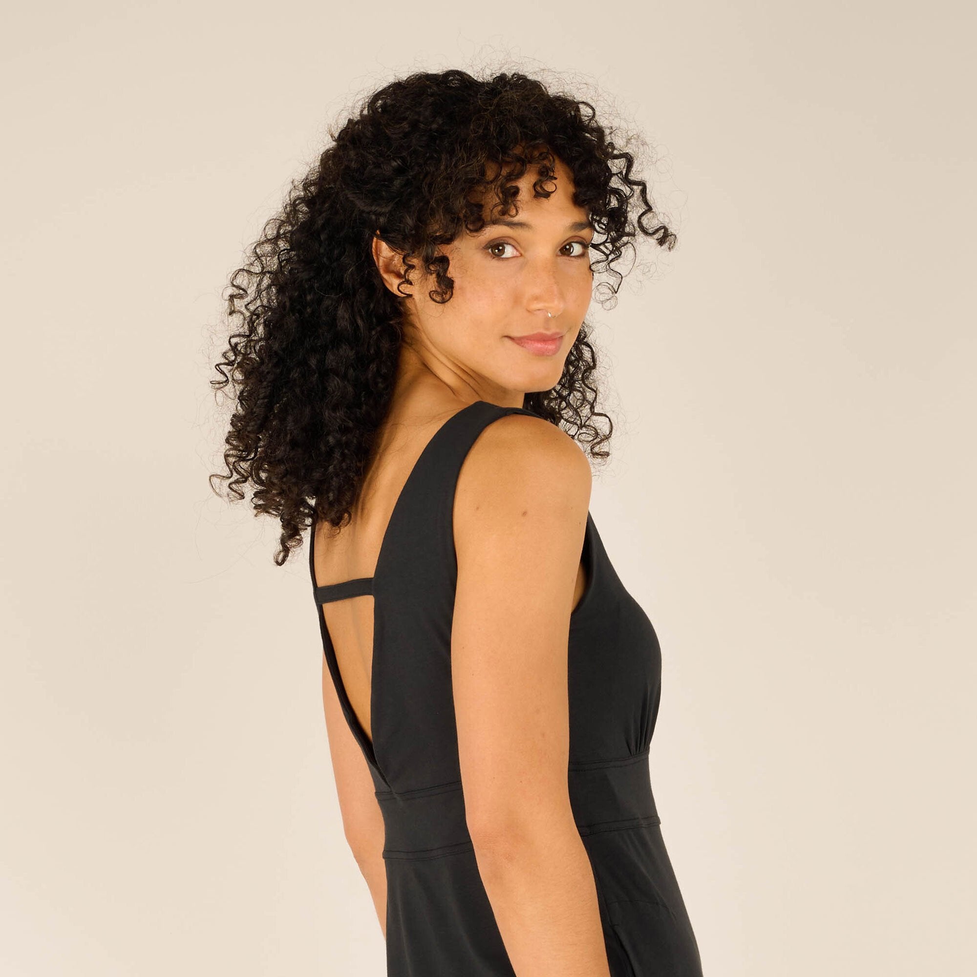 A detailed close-up of the model’s upper back, showing the elegant deep V-cut with the thin strap, along with the soft drape of the Sherpa Adventure Gear Neha Midi Dress in Blacks fabric. Her curly hair is slightly pulled back to emphasise the cutout.