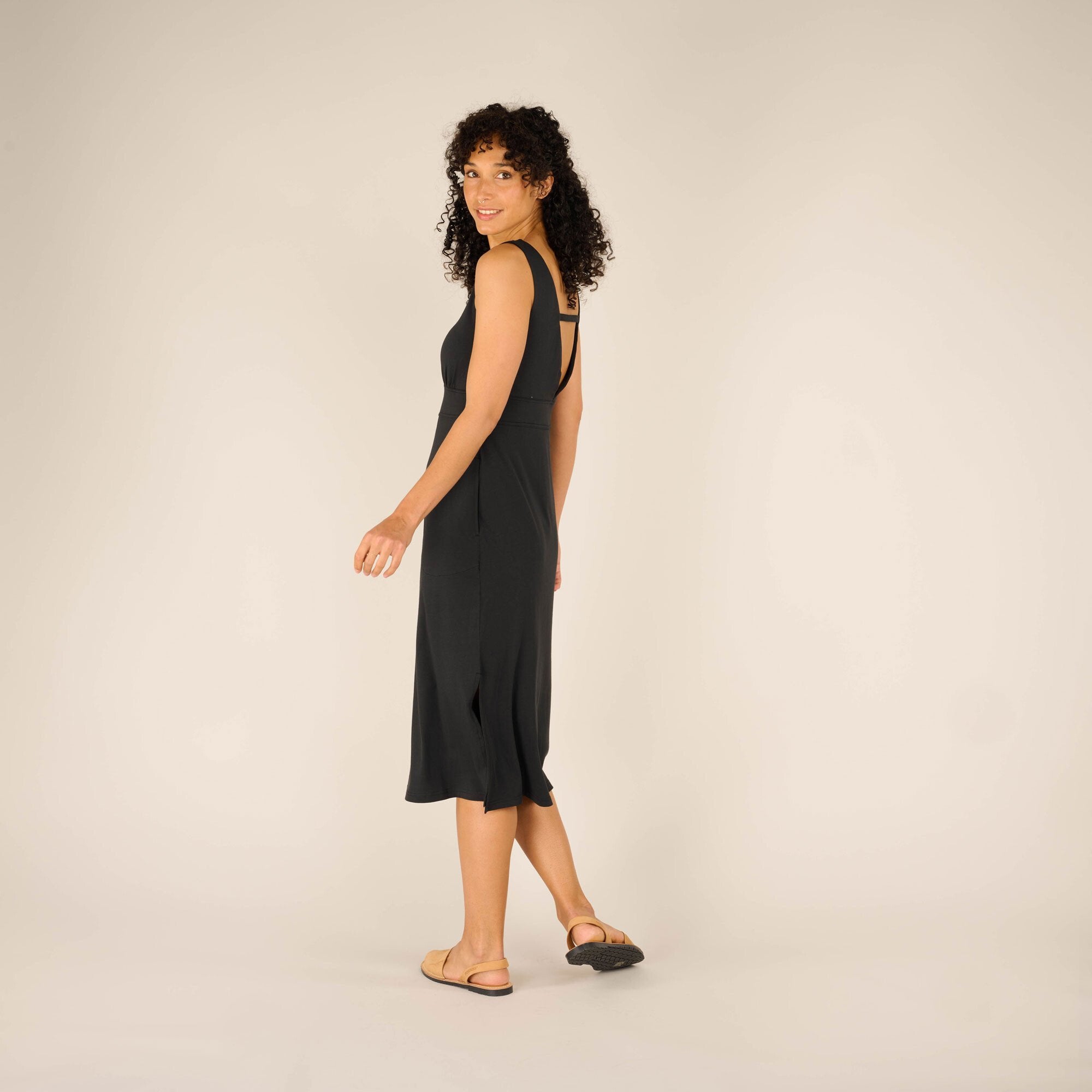 The model turns slightly with one foot stepping forward, glancing back towards the camera. The movement showcases the Sherpa Adventure Gear Neha Midi Dress in Black’s slight flare and side slit detail. Her tan sandals complement the sleek black fabric.