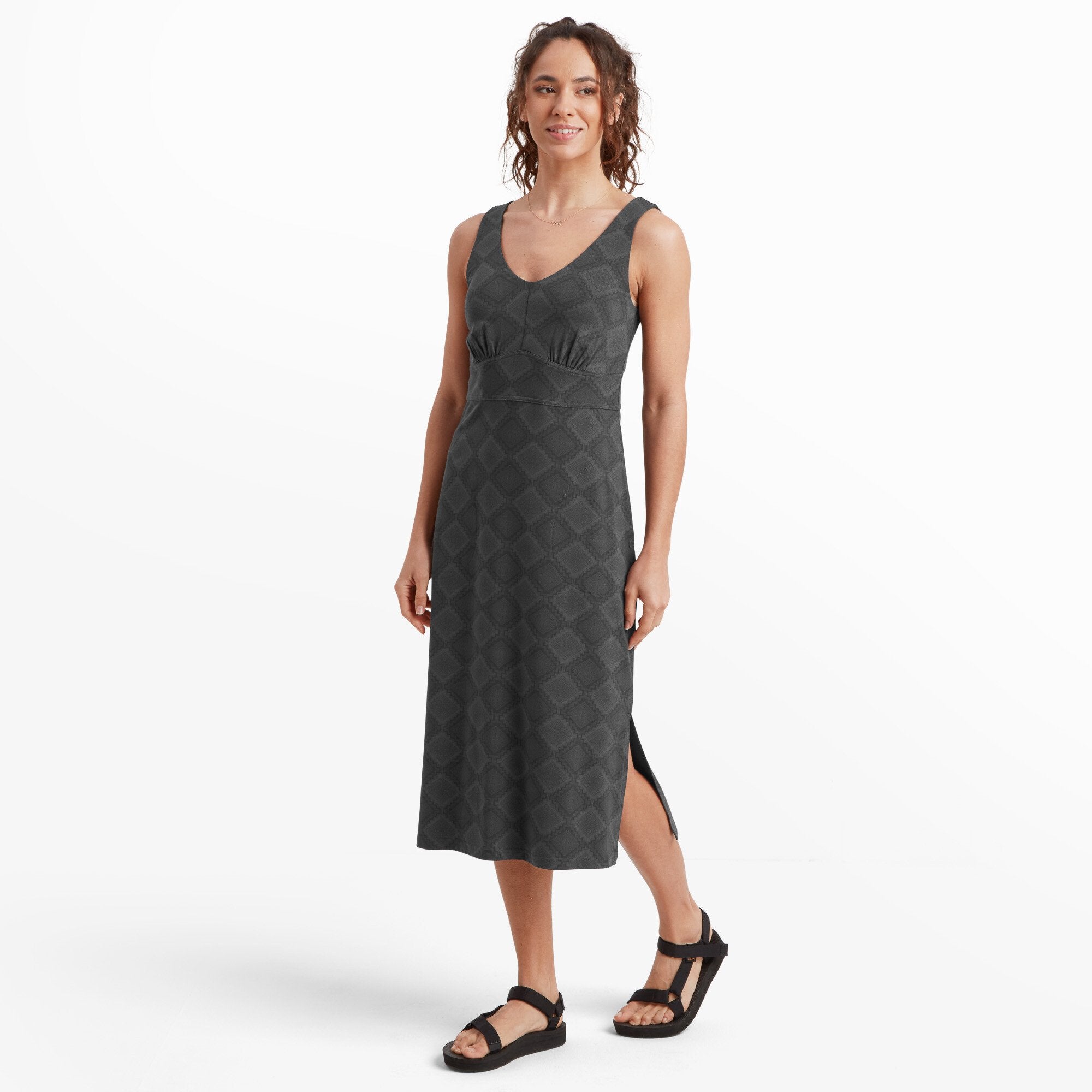 A woman standing confidently in a Sherpa Adventure Gear Neha Midi Dress in Black with a subtle diamond-shaped pattern. The dress has a fitted bodice with a V-neckline and a gathered waist, creating a flattering silhouette. She pairs the dress with simple black sandals, standing on a white background.