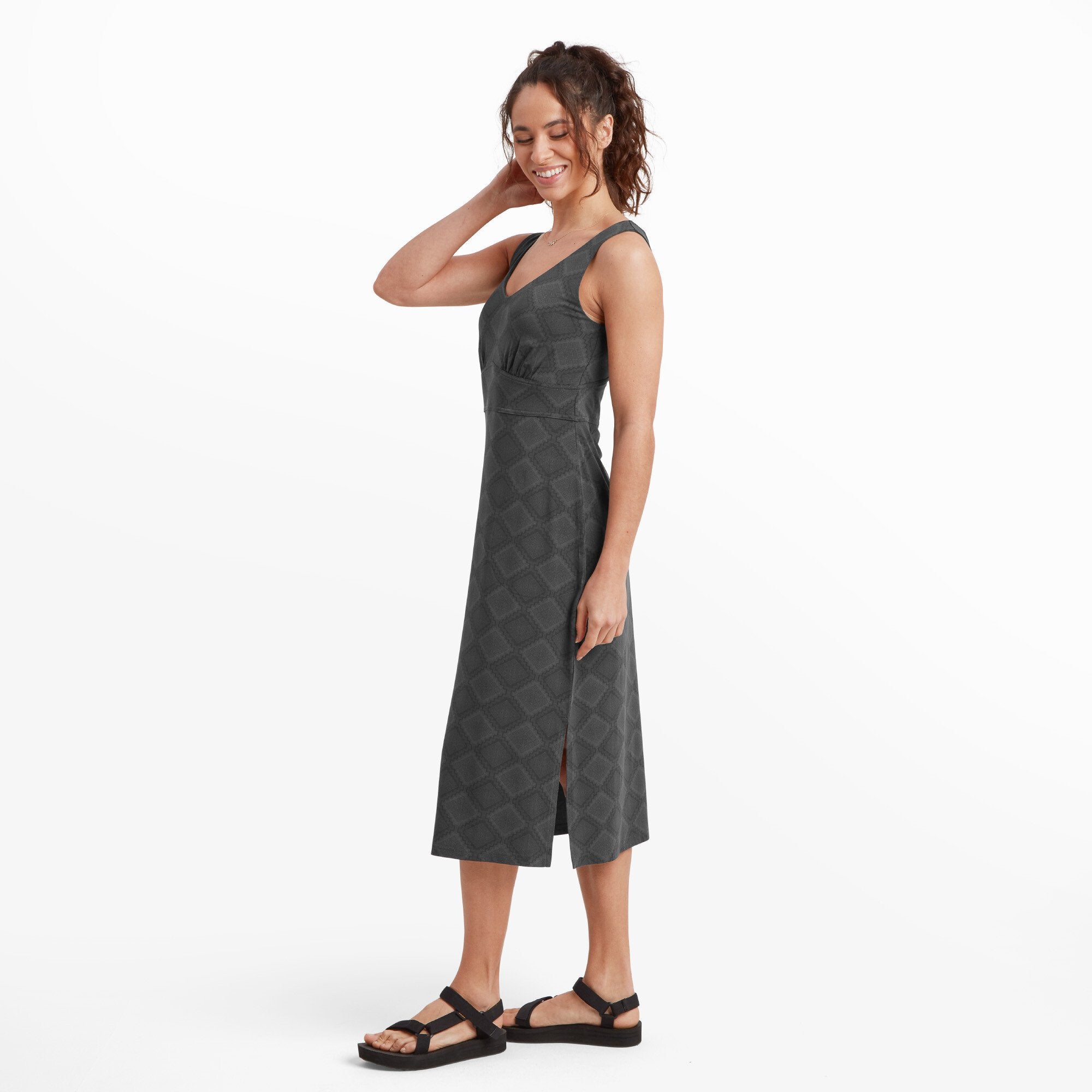 The model is shown from a three-quarters angle, emphasising the Sherpa Adventure Gear Neha Midi Dress in Black's structured fit along the waist and its elegant flow toward the hem. The side slit adds a touch of movement and sophistication. She looks down slightly with a gentle smile.